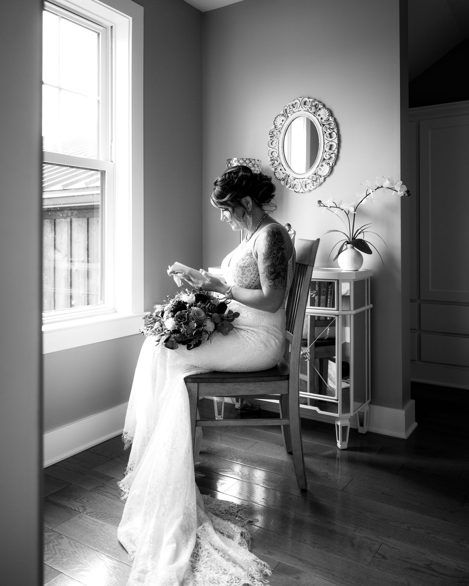 Black and white portrait of bride in lace gown sitting by window holding bouquet in elegant room.