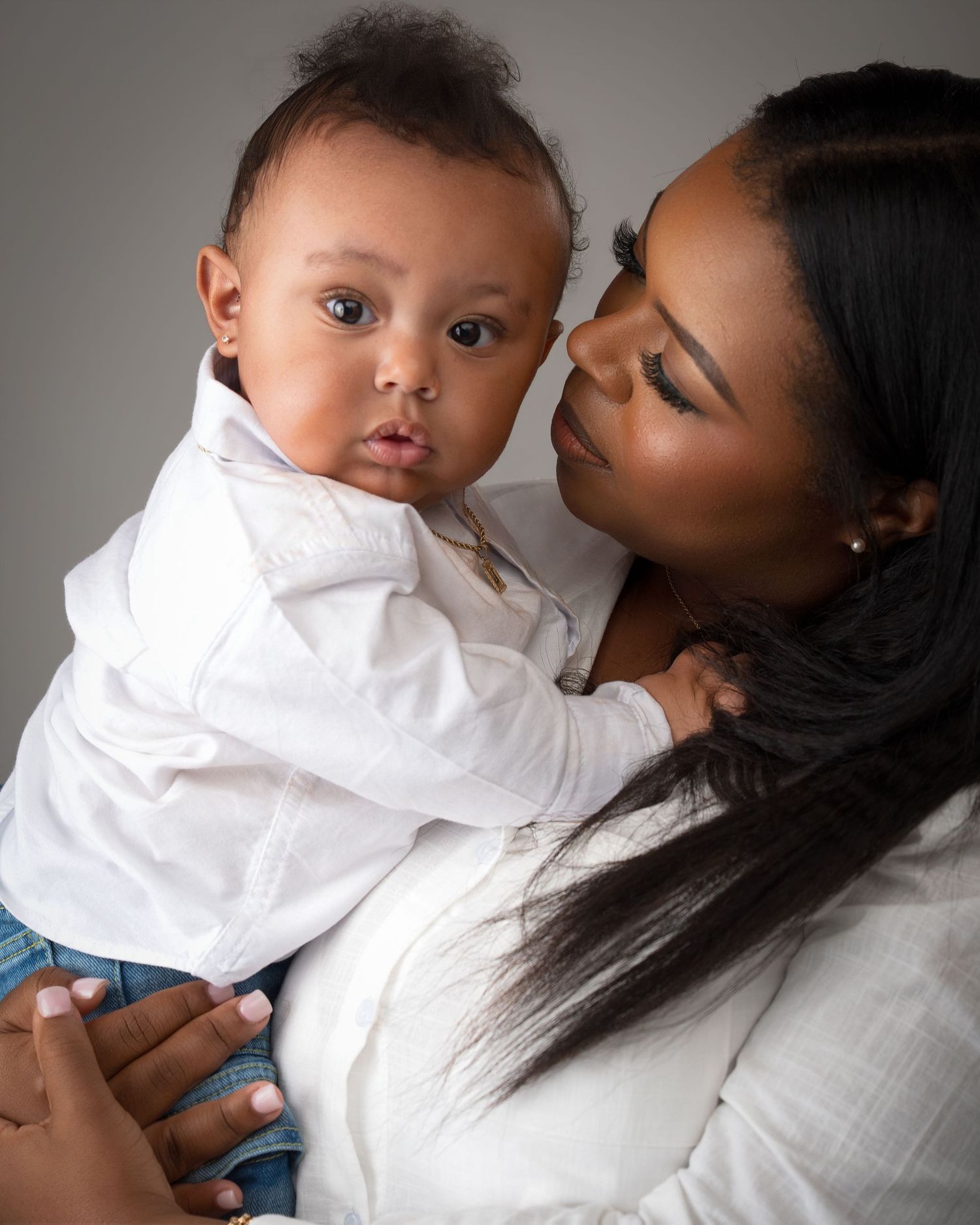 Mother gazes lovingly at alert baby boy in white shirt during professional studio portrait.