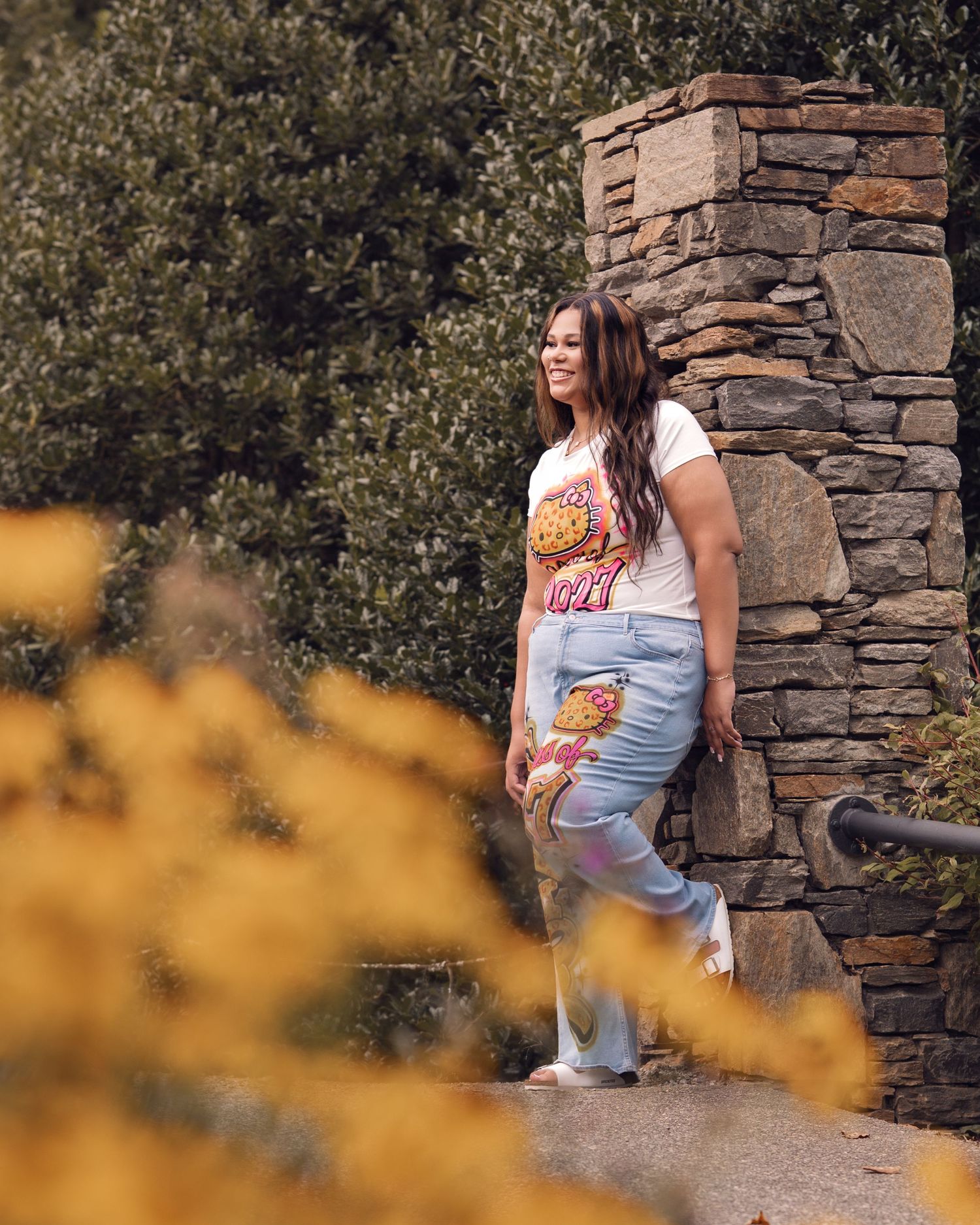Young woman in graphic tee and jeans leans against stone wall surrounded by fall foliage.