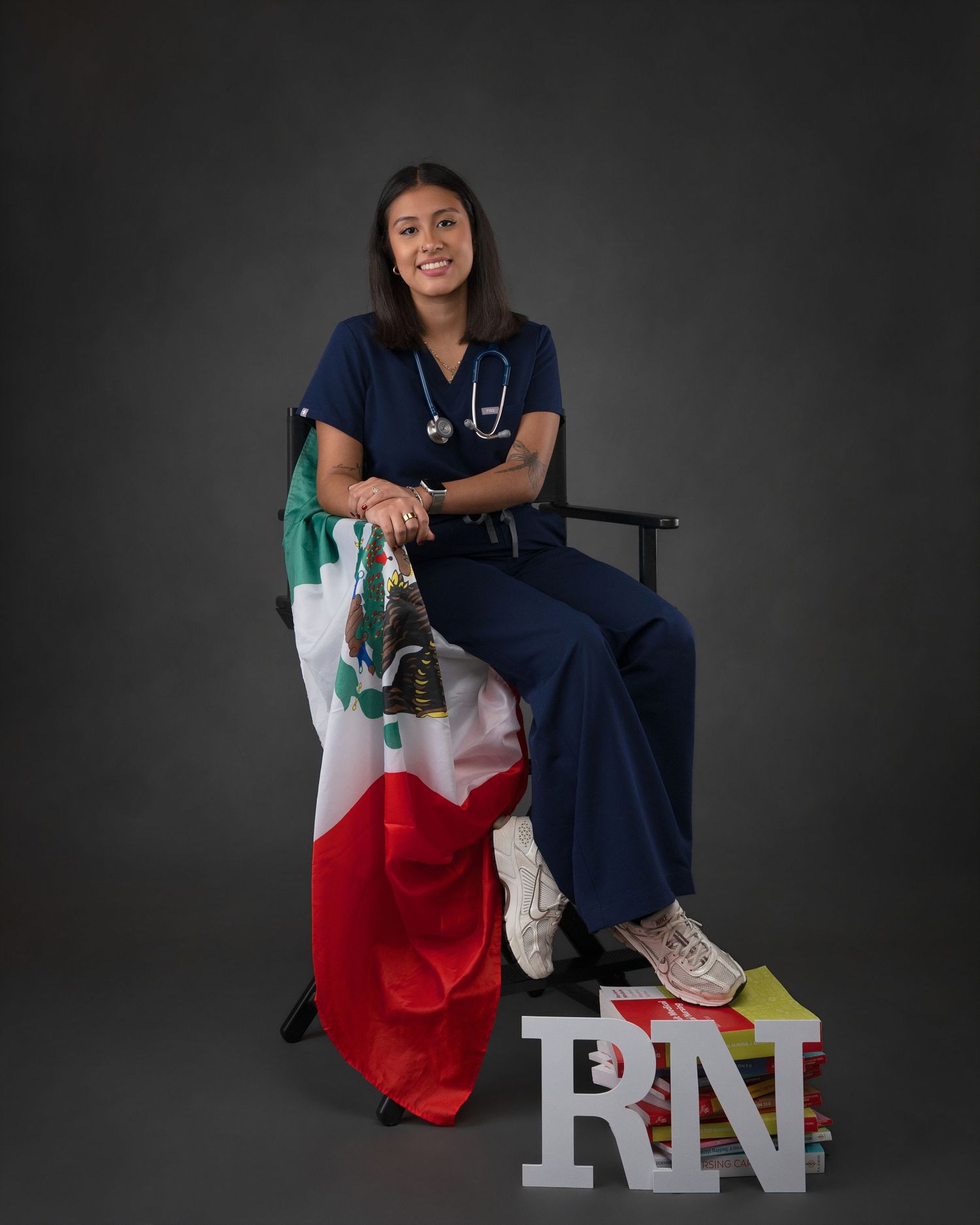 Female nurse in navy scrubs sits with a Mexican flag and RN letters in a graduation portrait.