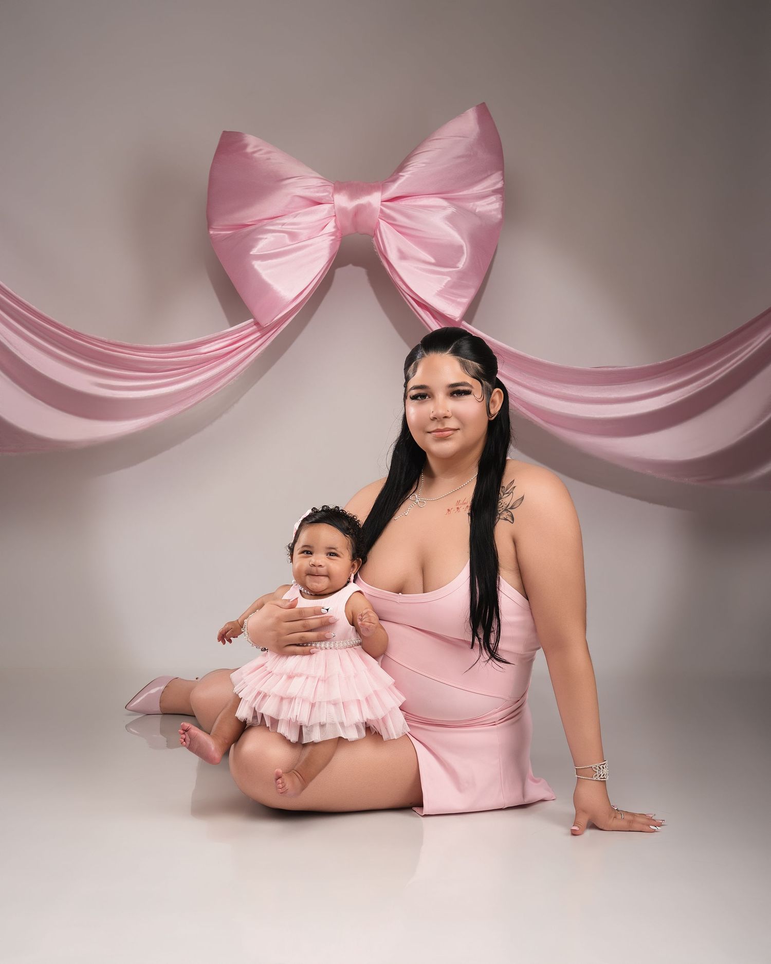 Mother and baby daughter pose together in matching pink outfits under large pink bow backdrop.