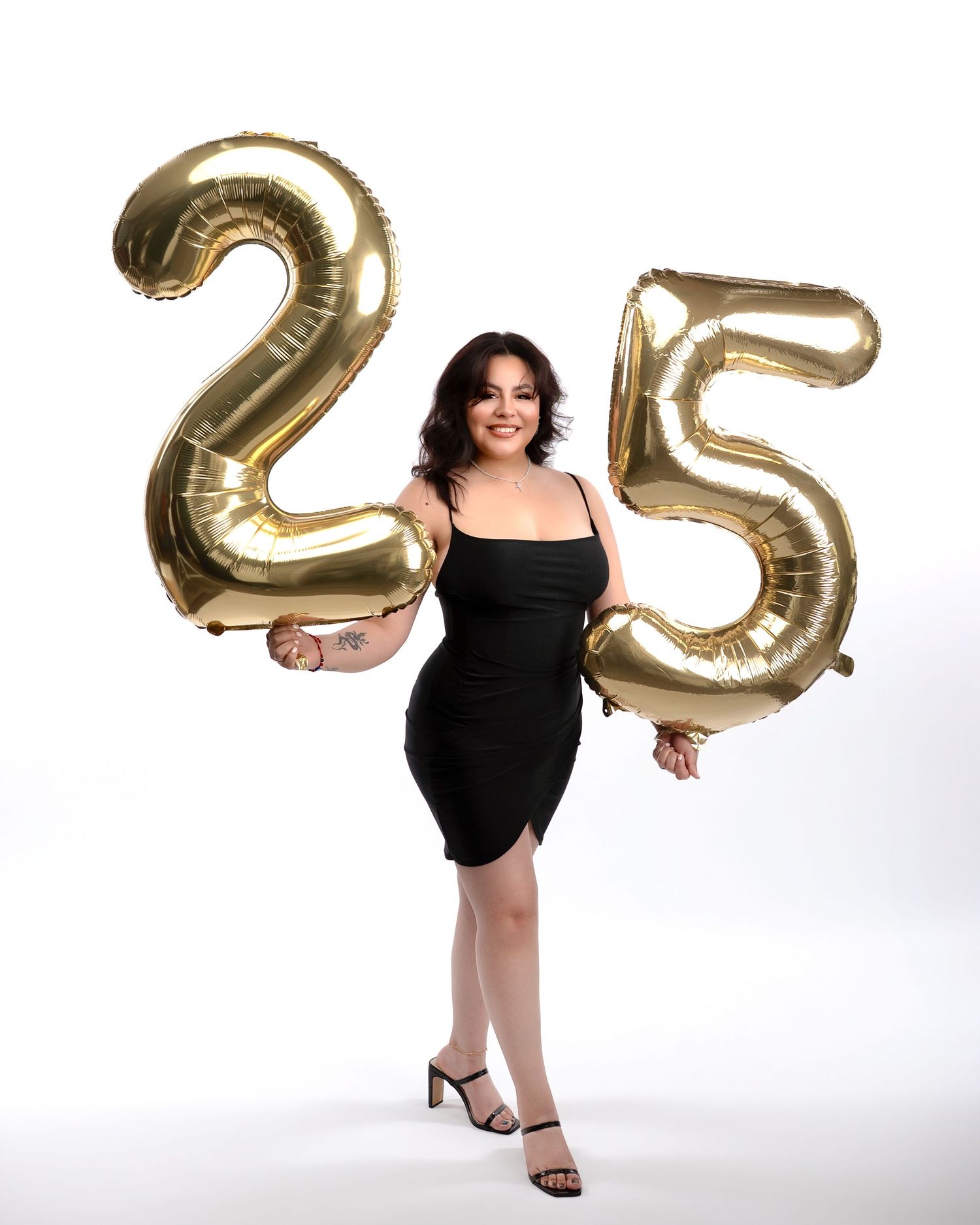 Woman in black dress smiles holding large gold number 25 balloons for her 25th birthday celebration.