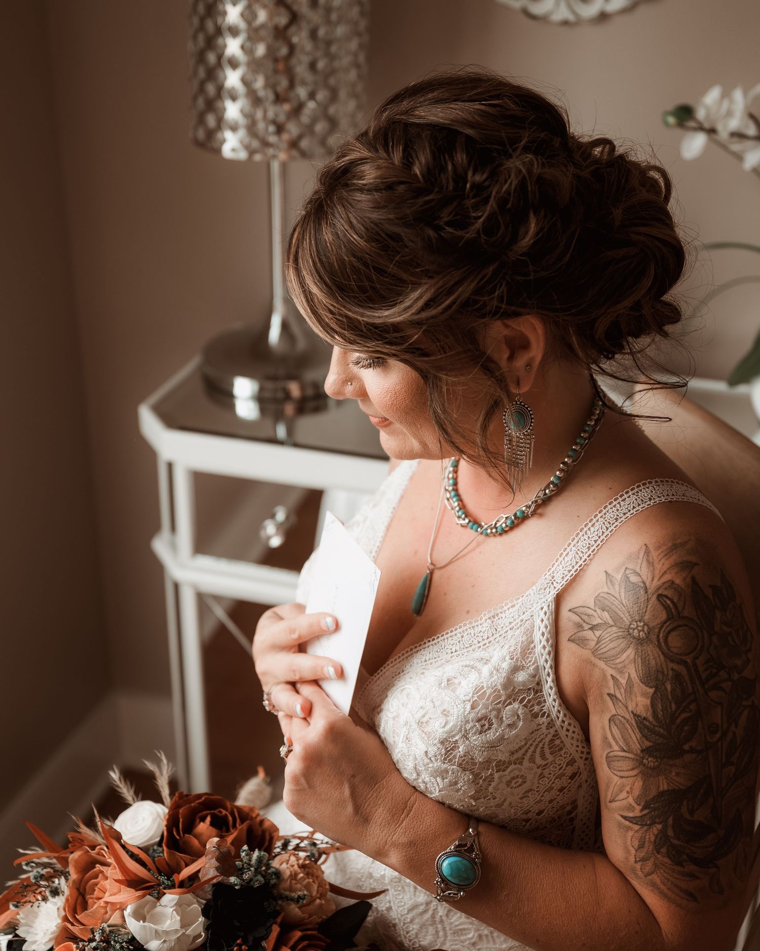Tattooed bride in lace gown reads a letter while holding a terracotta bouquet with turquoise jewelry.