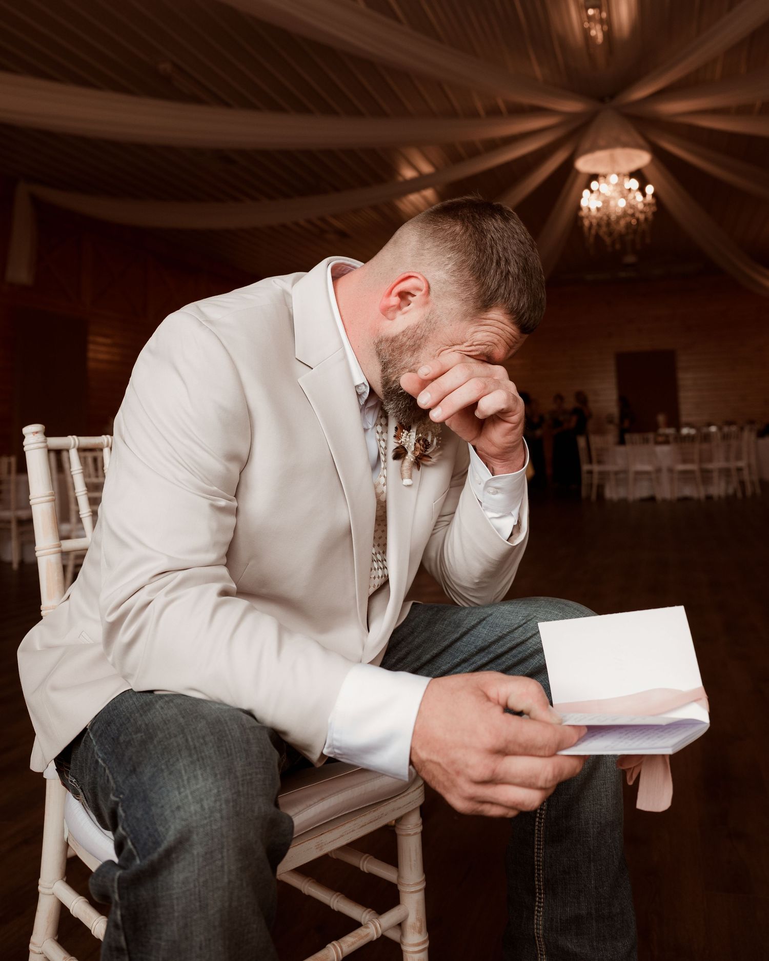 Emotional groom reads heartfelt letter from bride while seated in wedding venue ballroom.