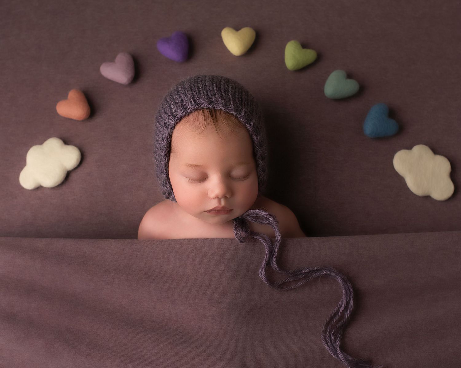 Sleeping newborn in purple knit bonnet surrounded by rainbow hearts and clouds on brown backdrop.