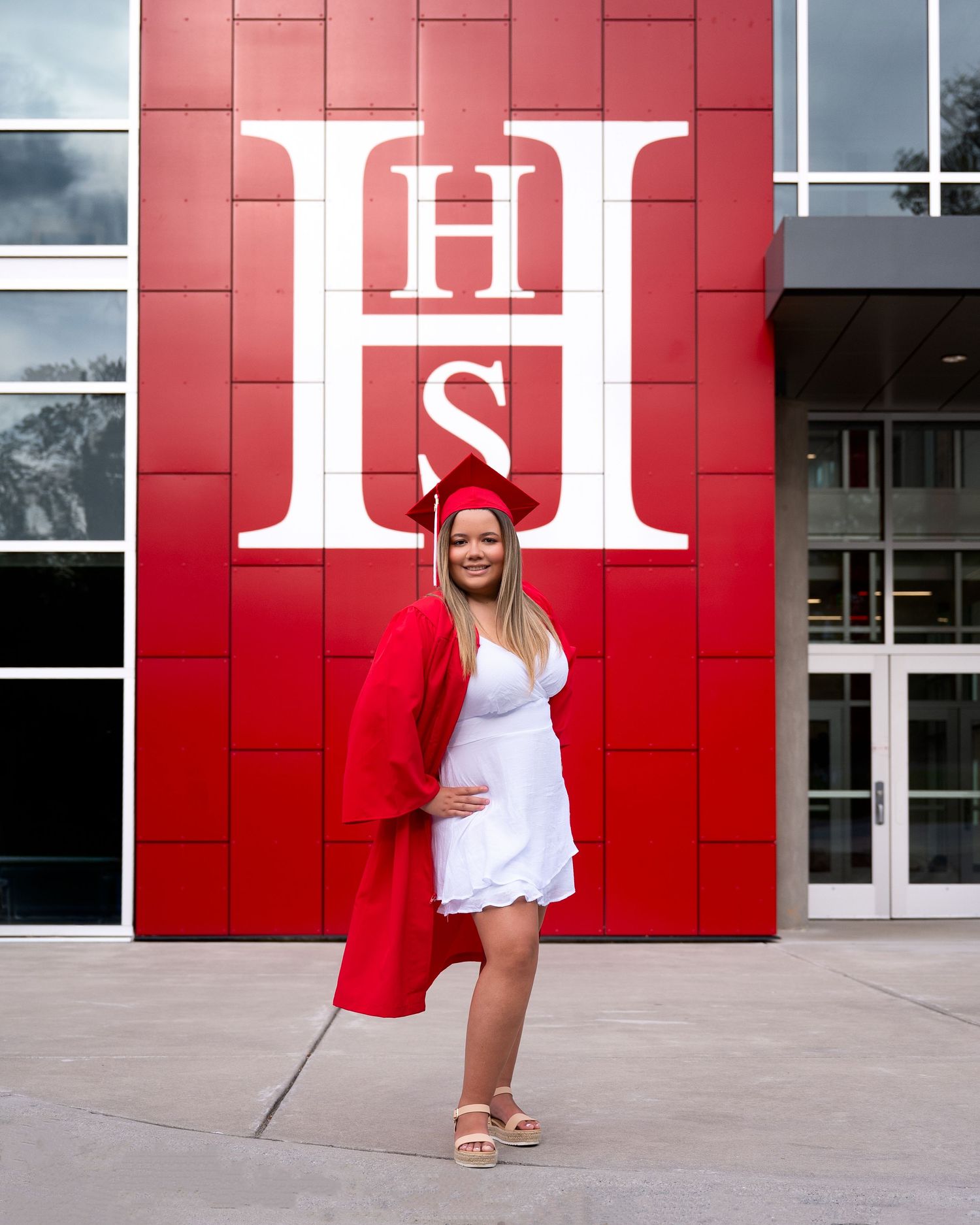 Pregnant graduate in red cap and gown poses in front of red school building with large H logo.