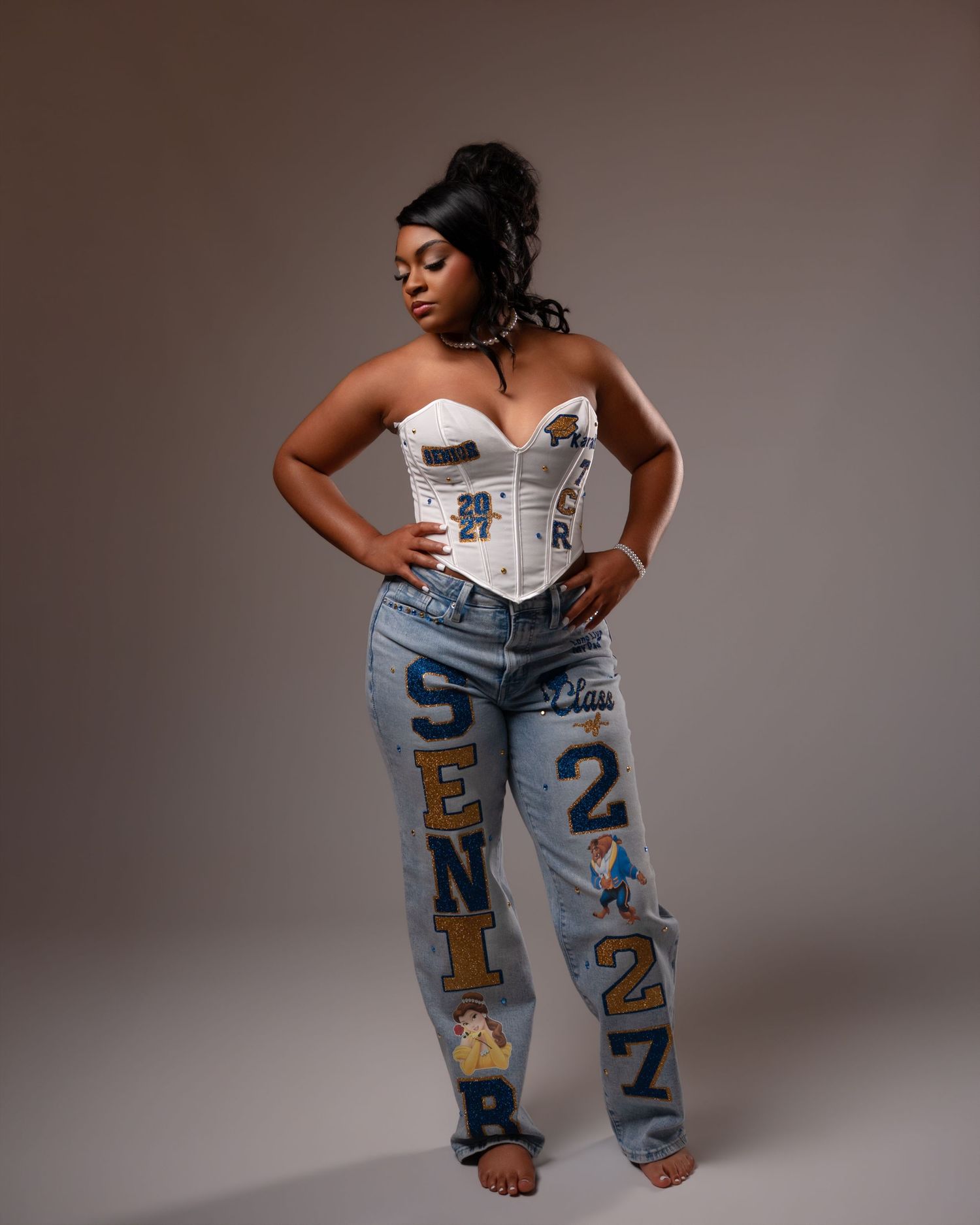Young woman in white corset top and custom SENIOR 2027 jeans poses in studio portrait.