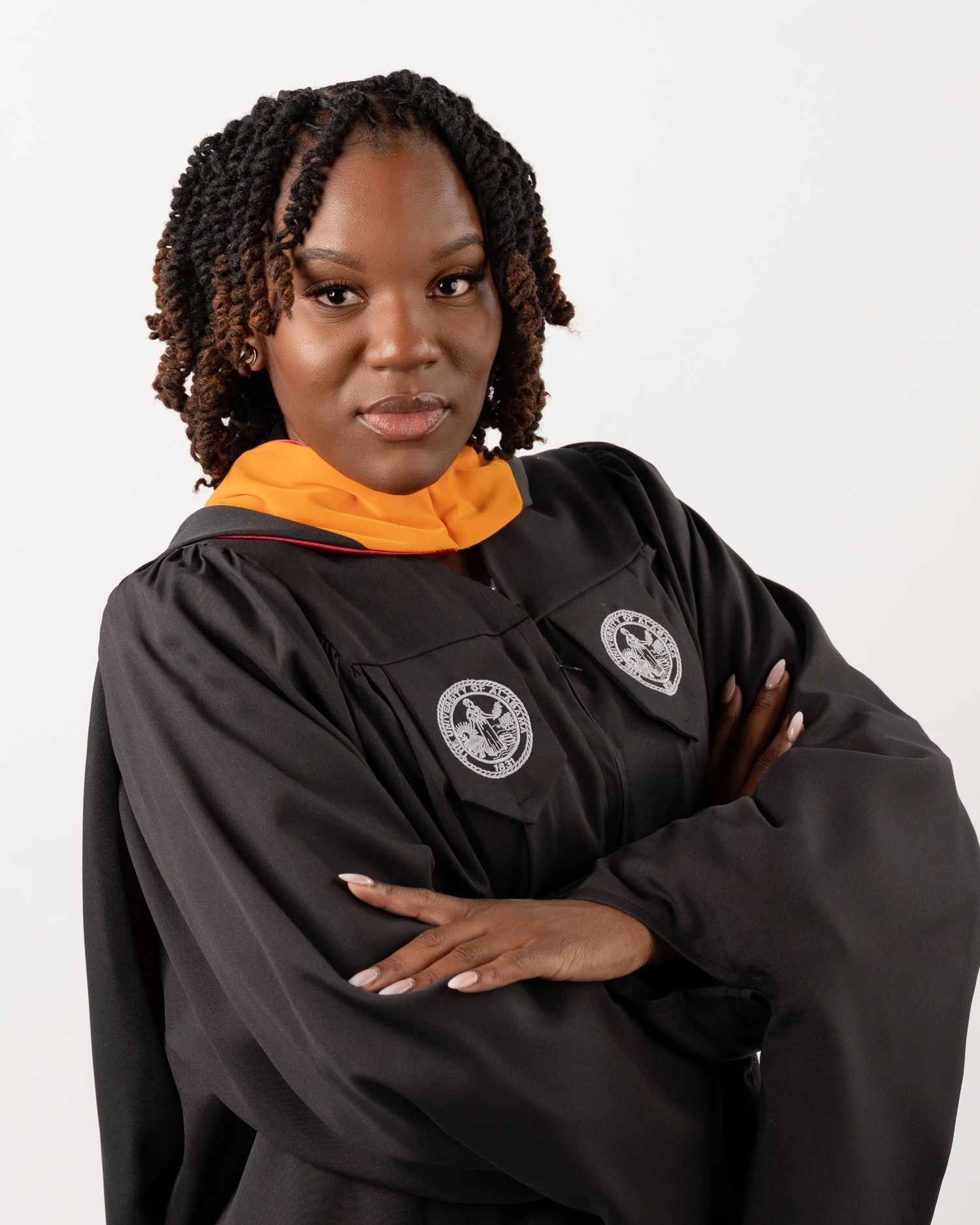 Graduate in black and orange doctoral regalia posing with arms crossed in studio portrait.