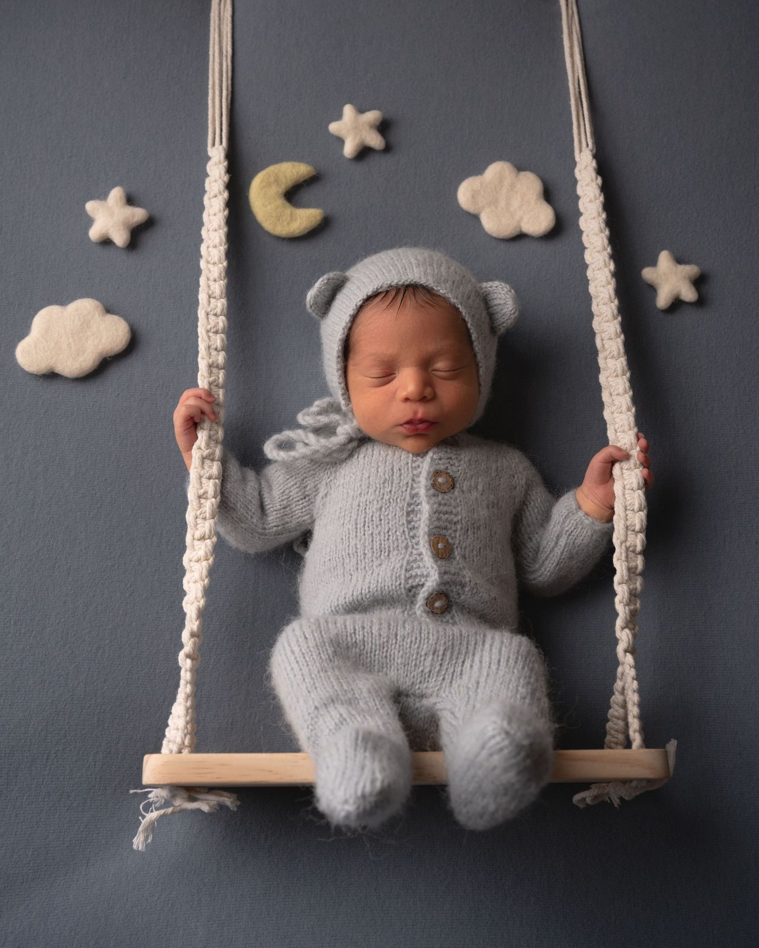 Sleeping newborn in gray knit bear outfit on wooden swing with star and cloud props on blue backdrop.