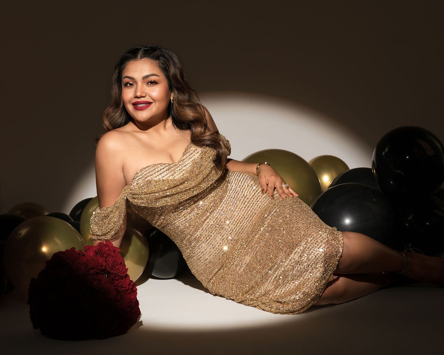 Woman in a gold sequin off-shoulder mini dress reclining on the floor beside black and gold balloons and a red rose bouquet.