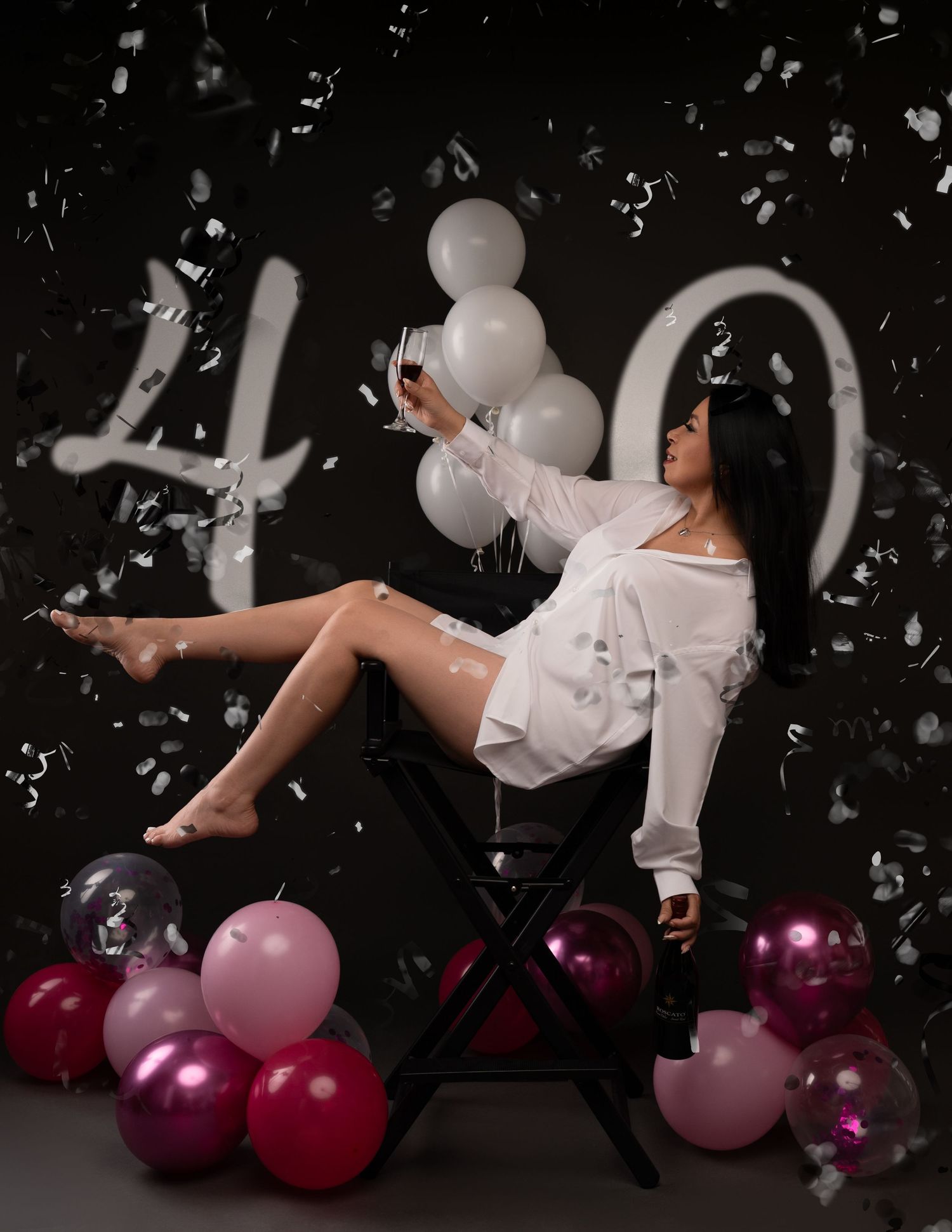 Woman in a white shirt reclines on a director's chair holding a wine glass, with balloons and a large 40 for a birthday photoshoot.