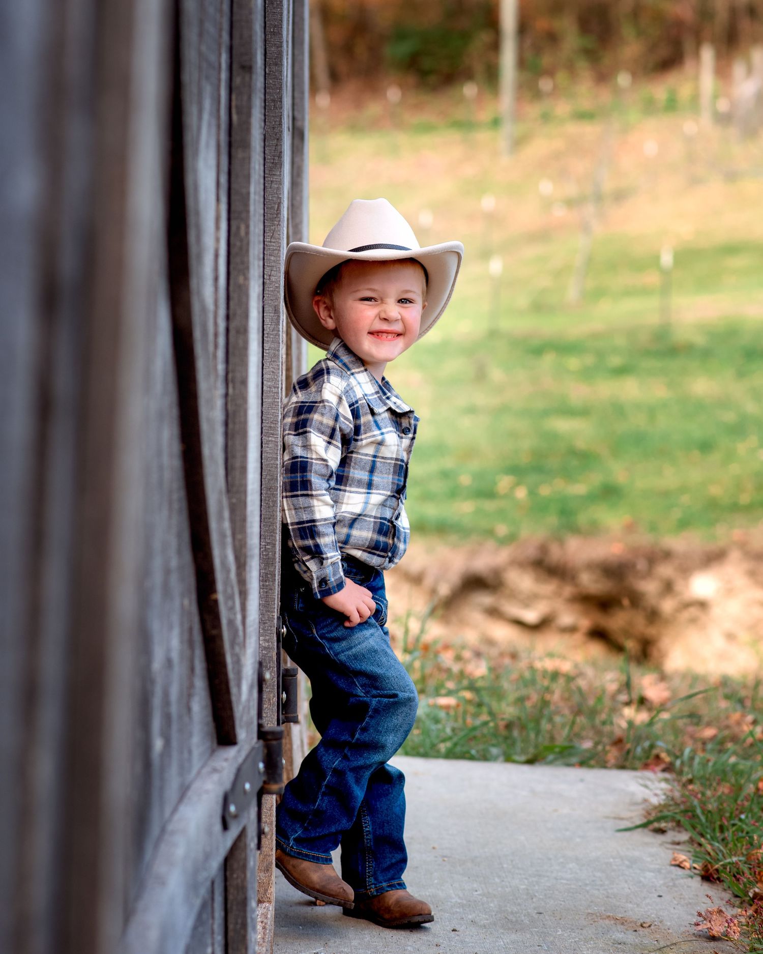 Sweet little boy in cowboy hat and boots posing by a rustic barn in jeans.