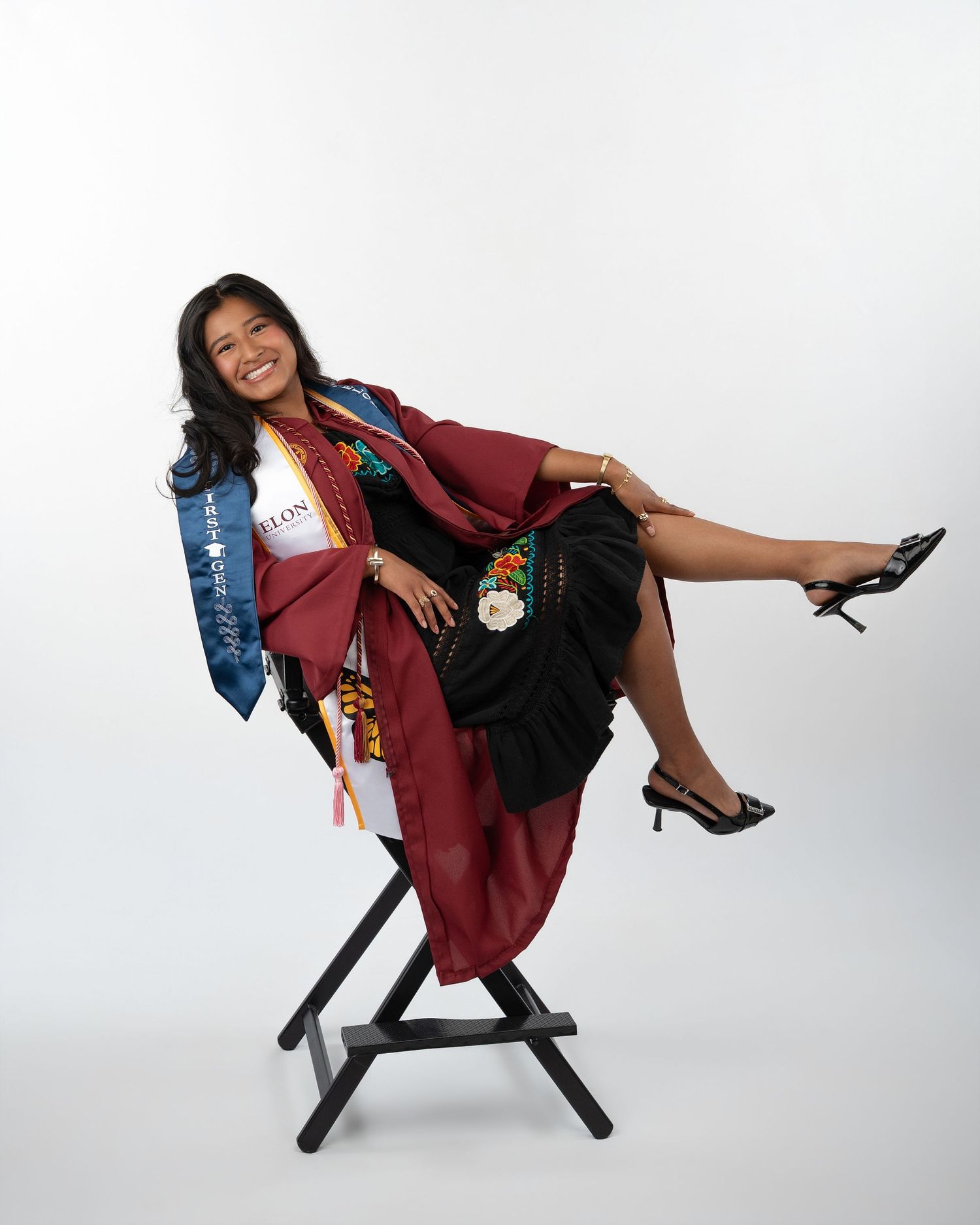Smiling female graduate in maroon gown with colorful sash poses playfully on chair in studio.