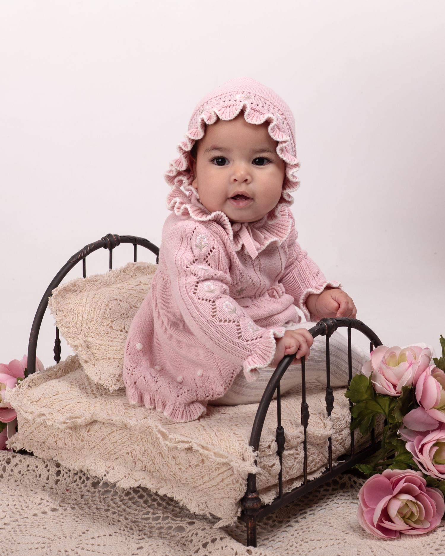 Sweet baby in pink knit dress and bonnet poses on vintage iron bed surrounded by pink roses.