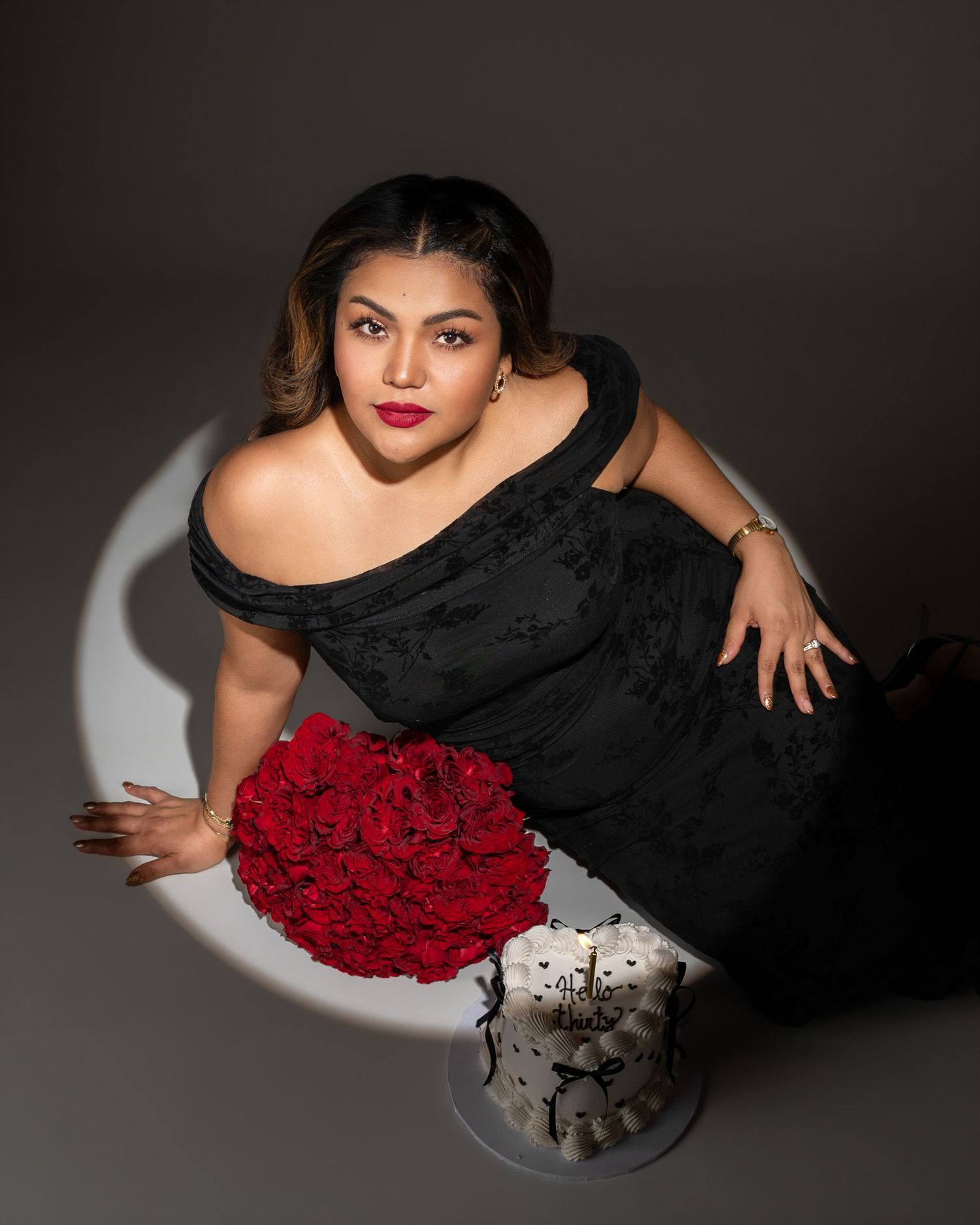 Woman in an off-shoulder black dress poses in a spotlight beside a bouquet of red roses and a small white birthday cake.