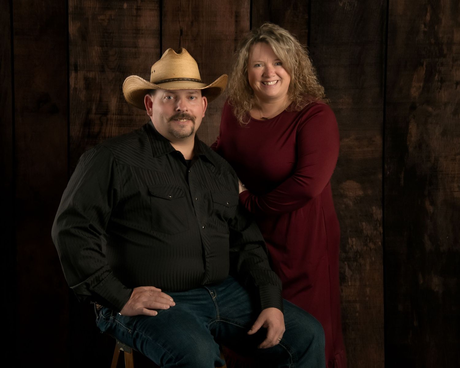 Couple in western attire with cowboy hats pose for portrait against rustic dark wood background.