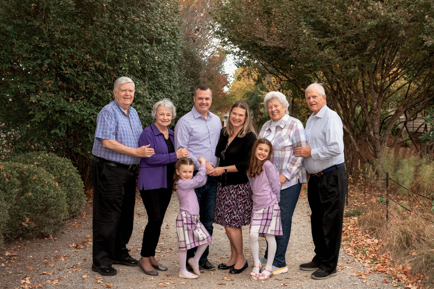Multi-generational family of eight poses outdoors on tree-lined path during autumn family portrait.