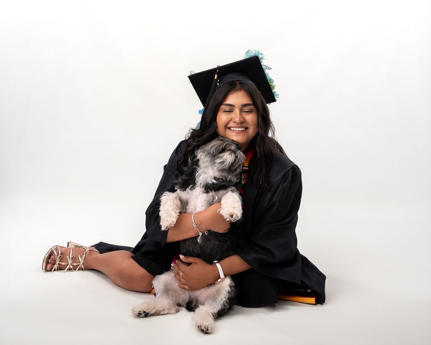 Smiling graduate in black cap and gown sits cross-legged hugging a fluffy dog during graduation photo session.