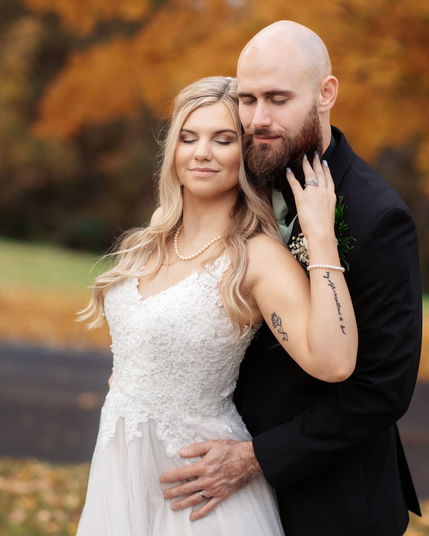 Bride and bearded groom embrace tenderly surrounded by vibrant autumn foliage.