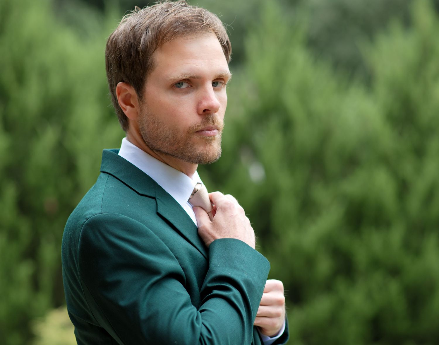 Handsome groom in a dark green suit adjusting his tie outdoors with evergreen trees in background.