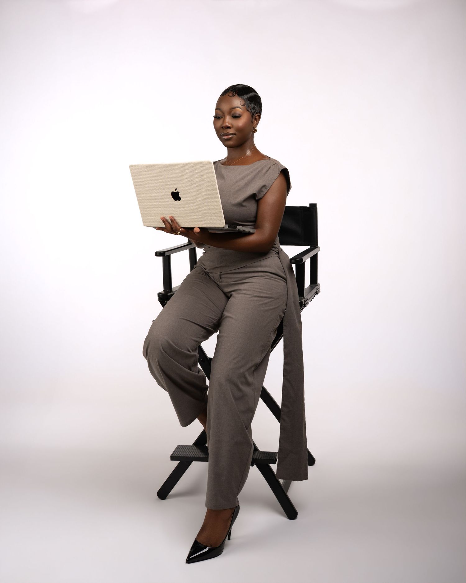 A professional woman in a gray jumpsuit sits in a director's chair holding an Apple MacBook laptop.