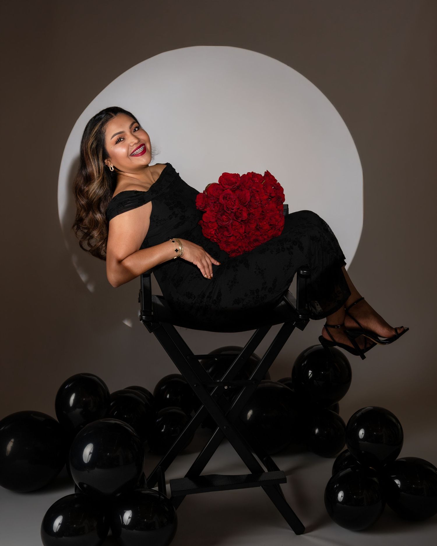 Woman in a black off-shoulder dress reclines in a director's chair holding red roses, surrounded by black balloons.