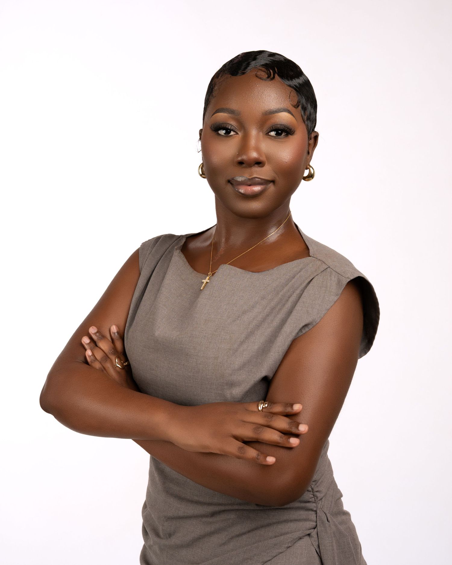 Confident woman with short styled hair, gold earrings and a cross necklace, wearing a taupe sleeveless top and posing with arms crossed.