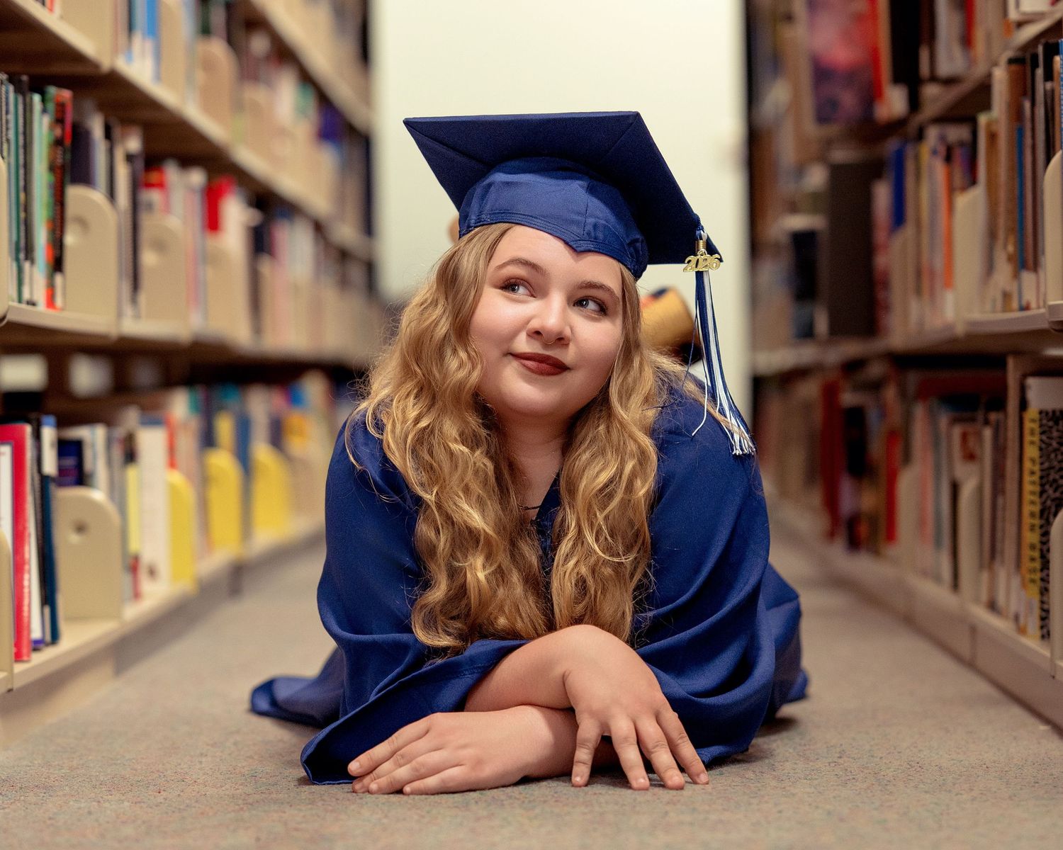 Blonde girl in blue graduation cap and gown lying on library floor gazing upward between bookshelves.