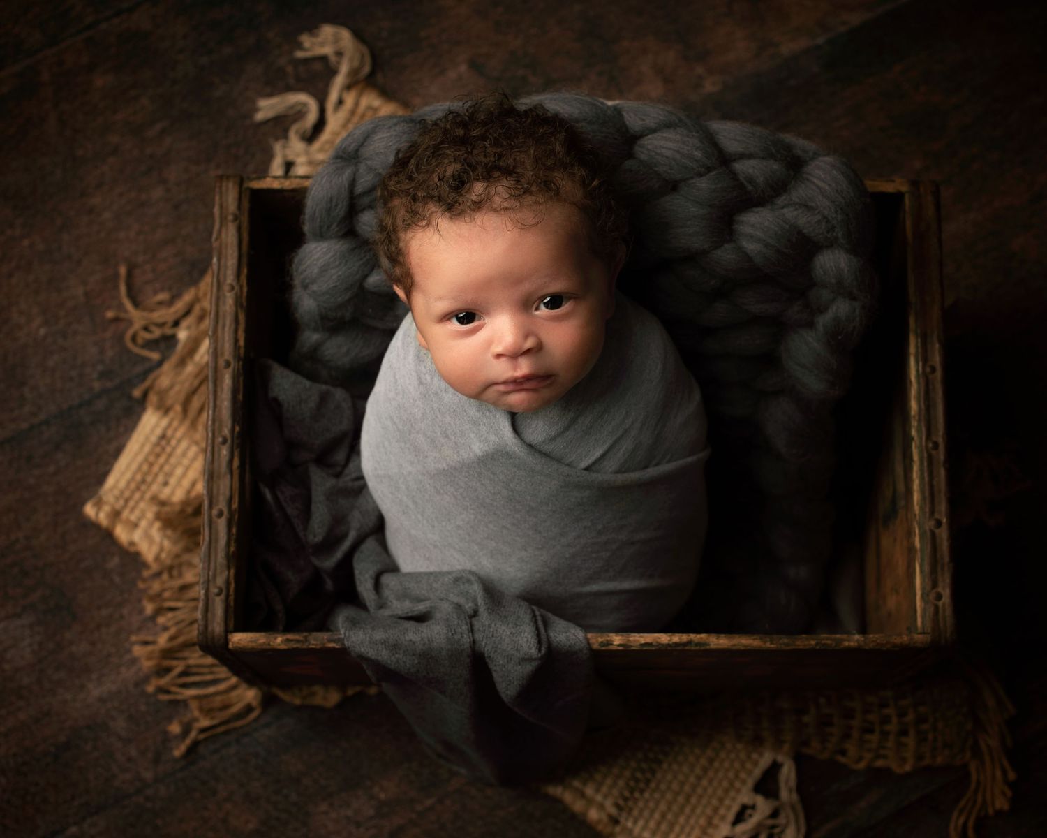 Alert newborn baby wrapped in gray swaddle nestled in a rustic wooden crate with yarn.