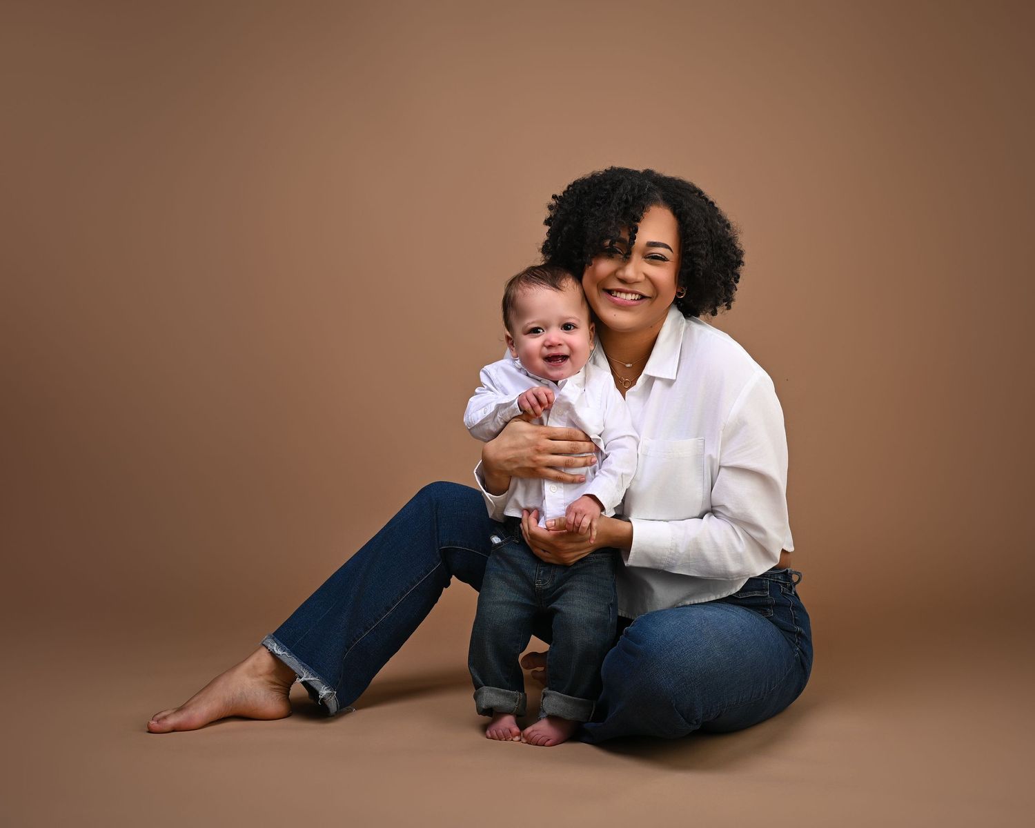 A curly-haired mother in white shirt sits smiling while holding her laughing baby on tan backdrop.