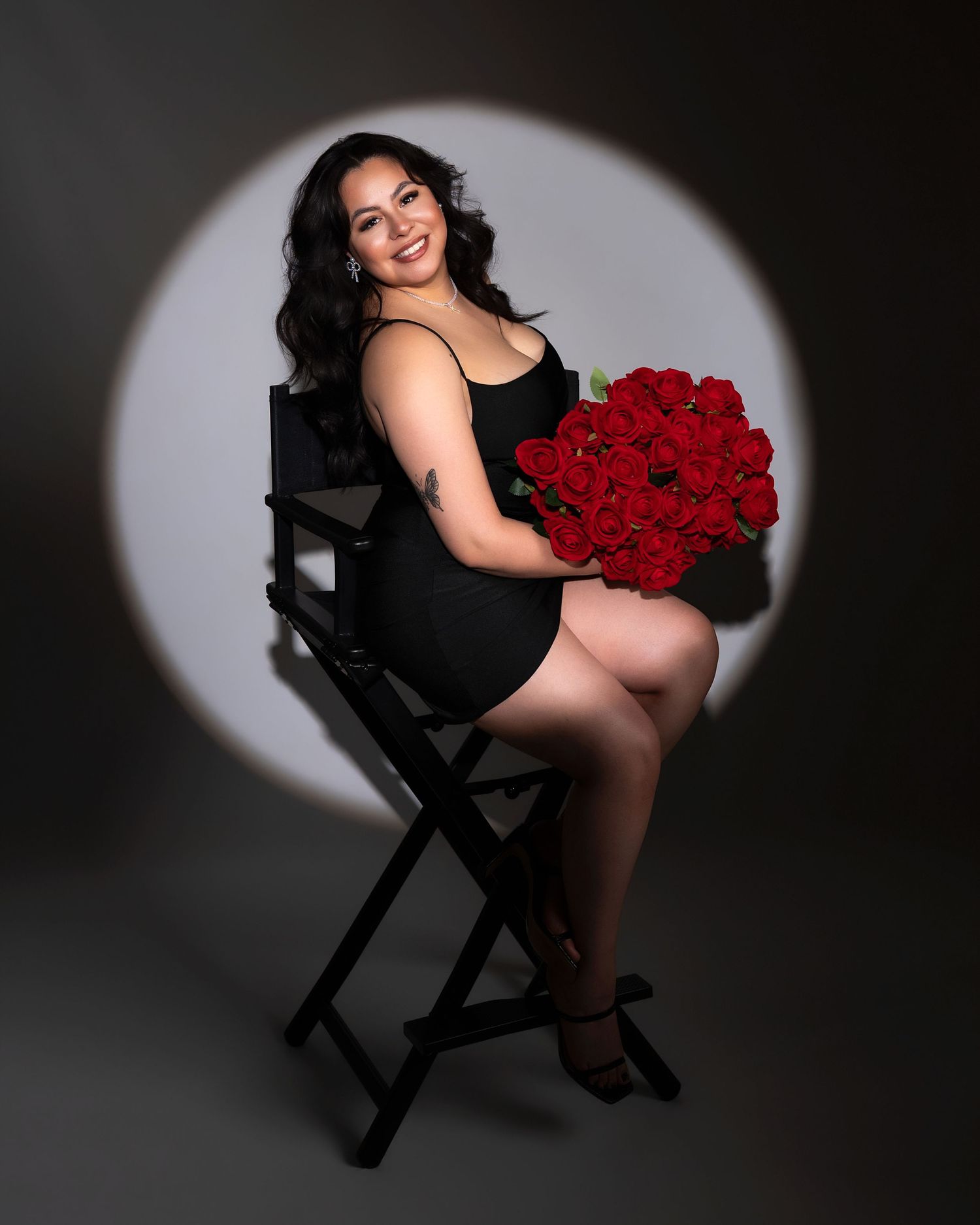 Woman in black dress sitting on director chair holding red roses bouquet in spotlight studio.