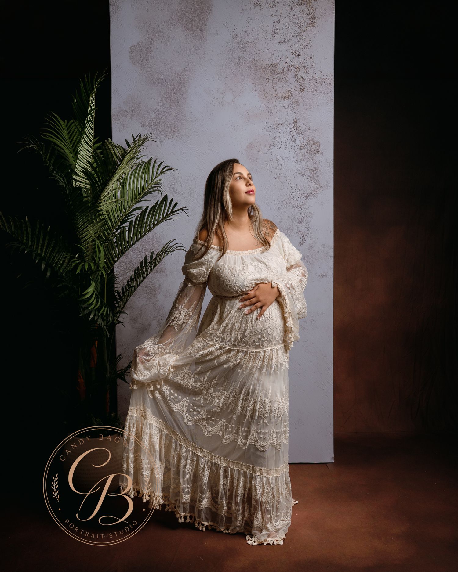 Pregnant woman in elegant white lace gown poses gracefully in a studio with a green plant and neutral backdrop.