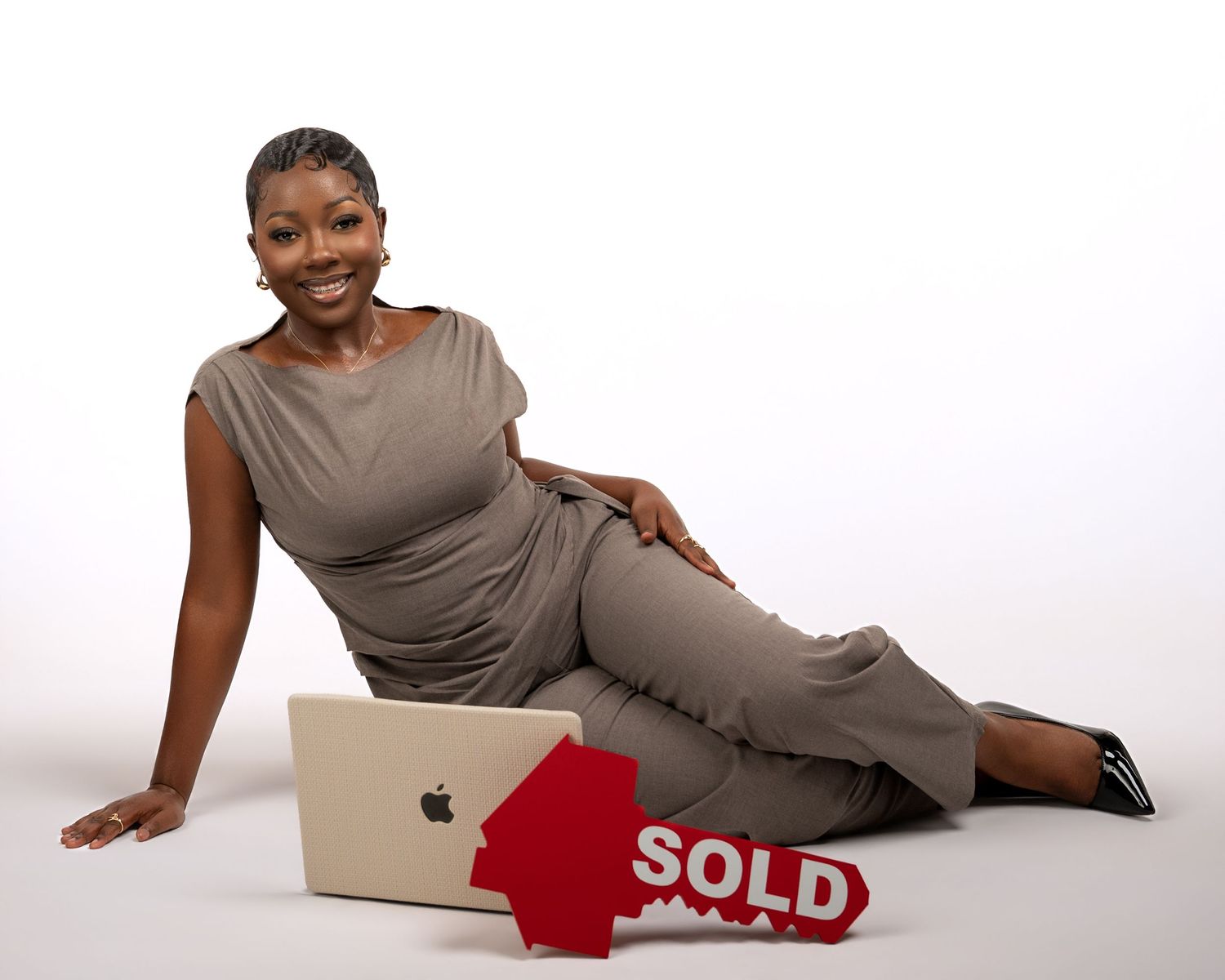 Woman in gray dress poses with Apple laptop and red SOLD real estate sign on white background.