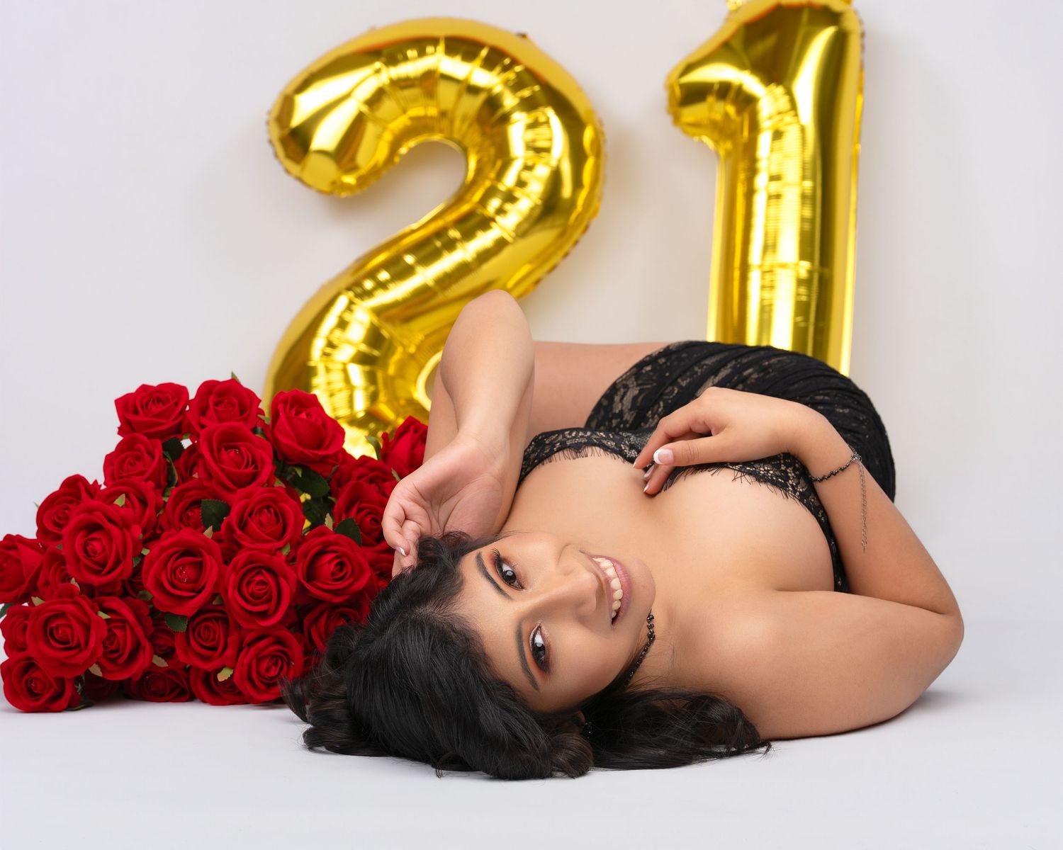 Woman in black lace lingerie lying on a white floor beside a bouquet of red roses and gold 21 balloons for a birthday photoshoot.