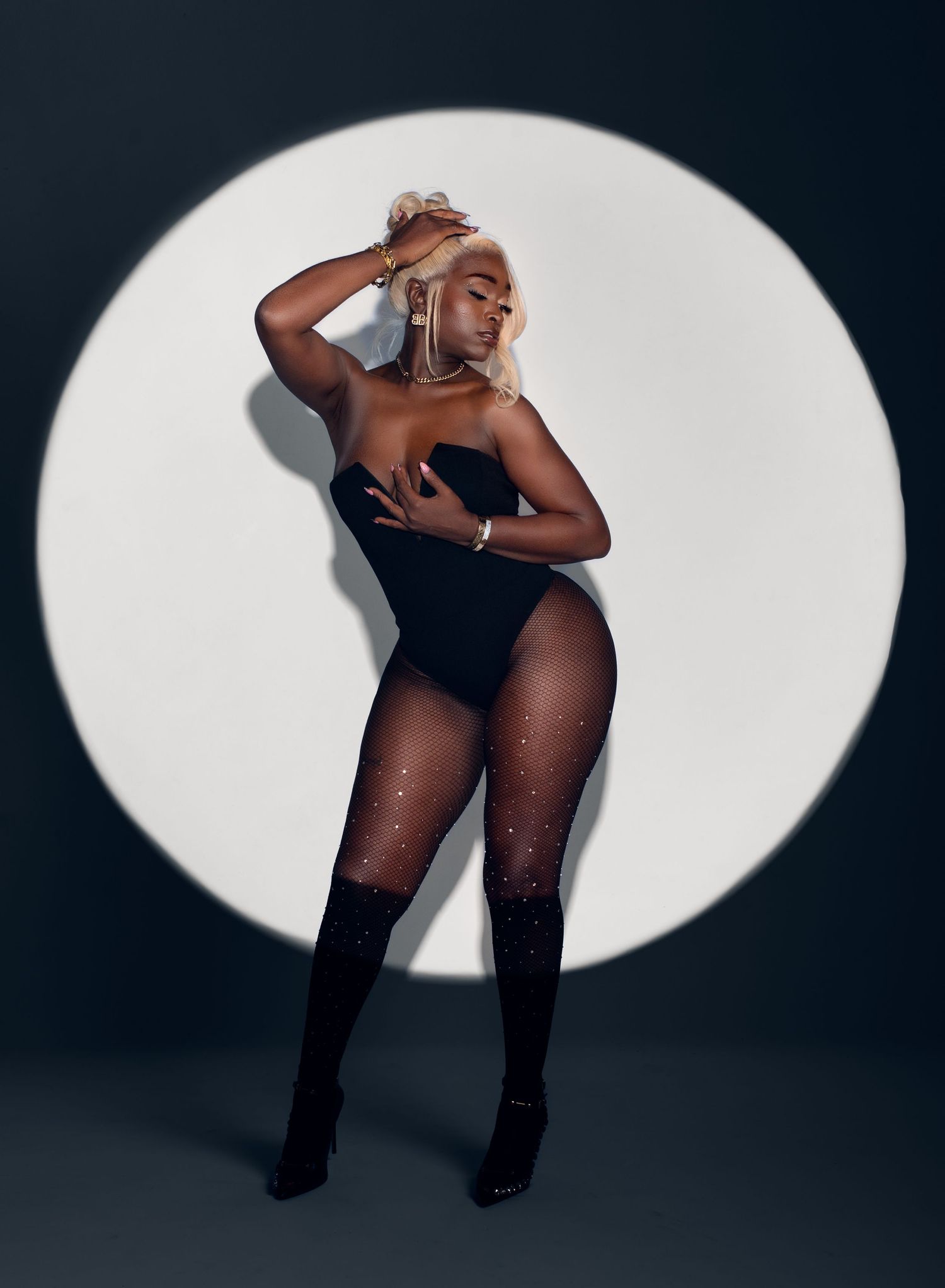 Woman with platinum blonde hair posing in a black strapless bodysuit and sparkling stockings against a glowing white circular spotlight.