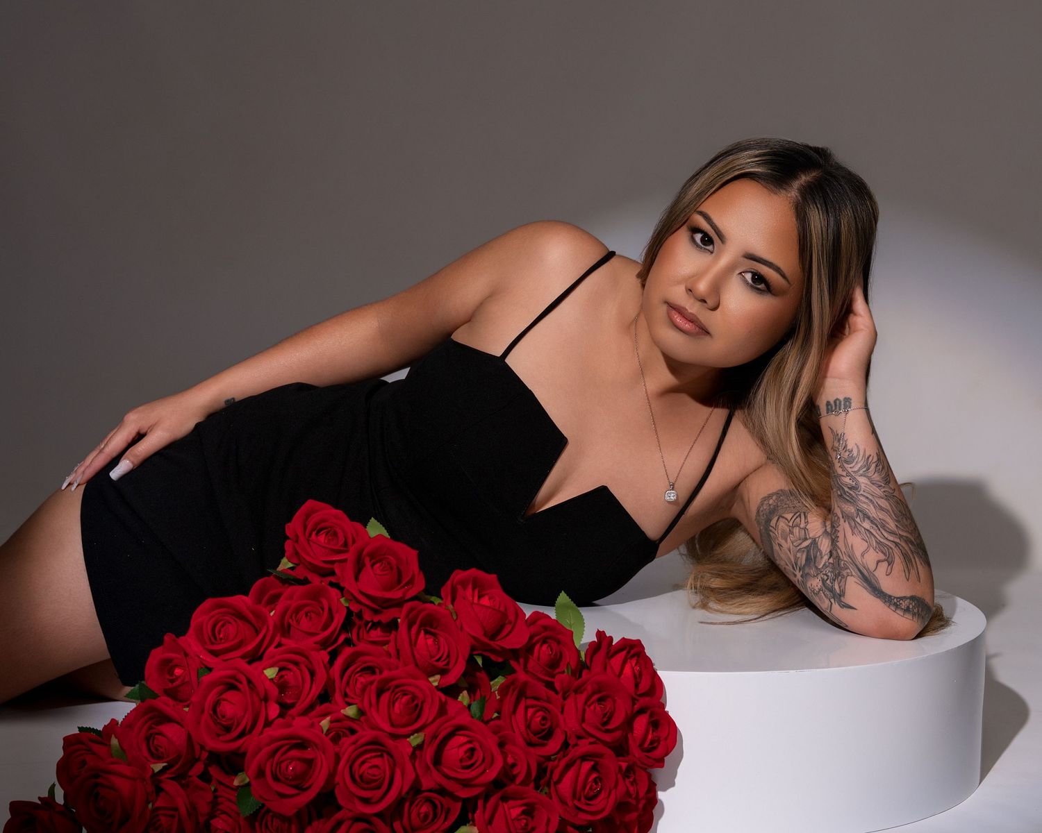 Woman in a black spaghetti-strap dress reclines on a white pedestal beside a bouquet of red roses in a moody studio portrait.