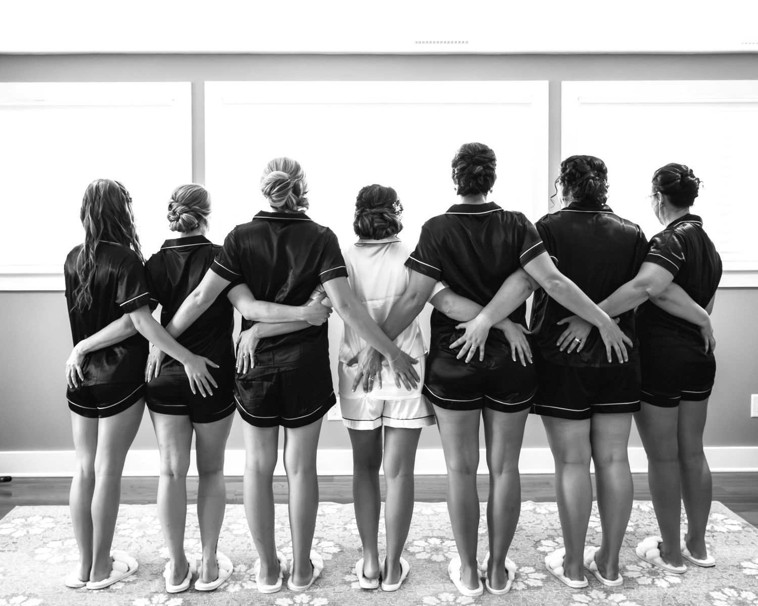Black and white photo of bridesmaids in matching robes standing arm-in-arm before a window.