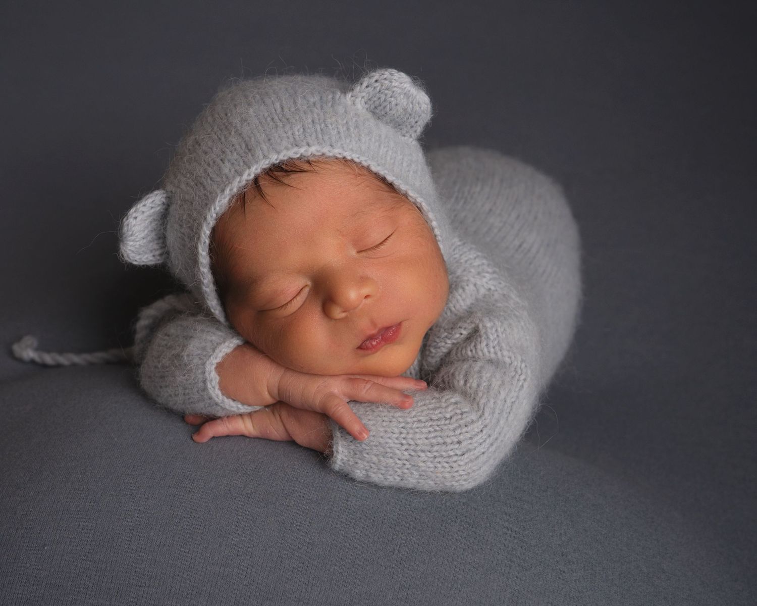 Adorable newborn baby sleeping in a gray knit bear outfit and bonnet on a gray backdrop.