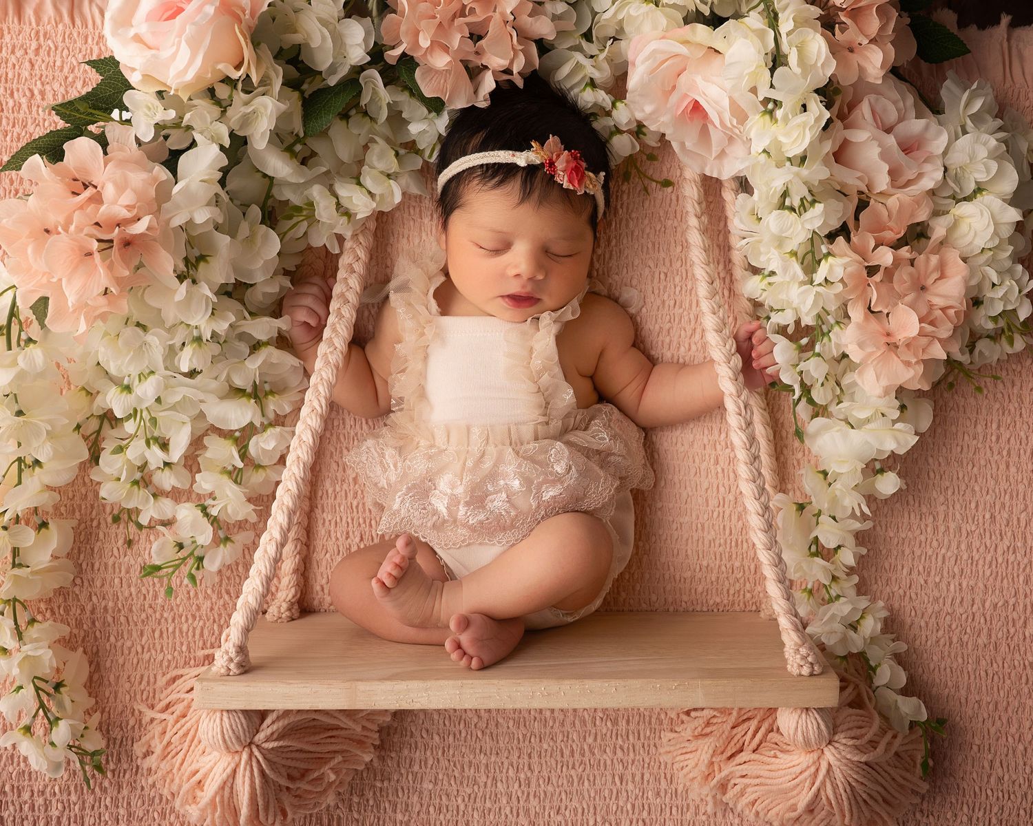 Sleeping newborn baby girl in lace romper posed on a wooden swing surrounded by pink flowers.