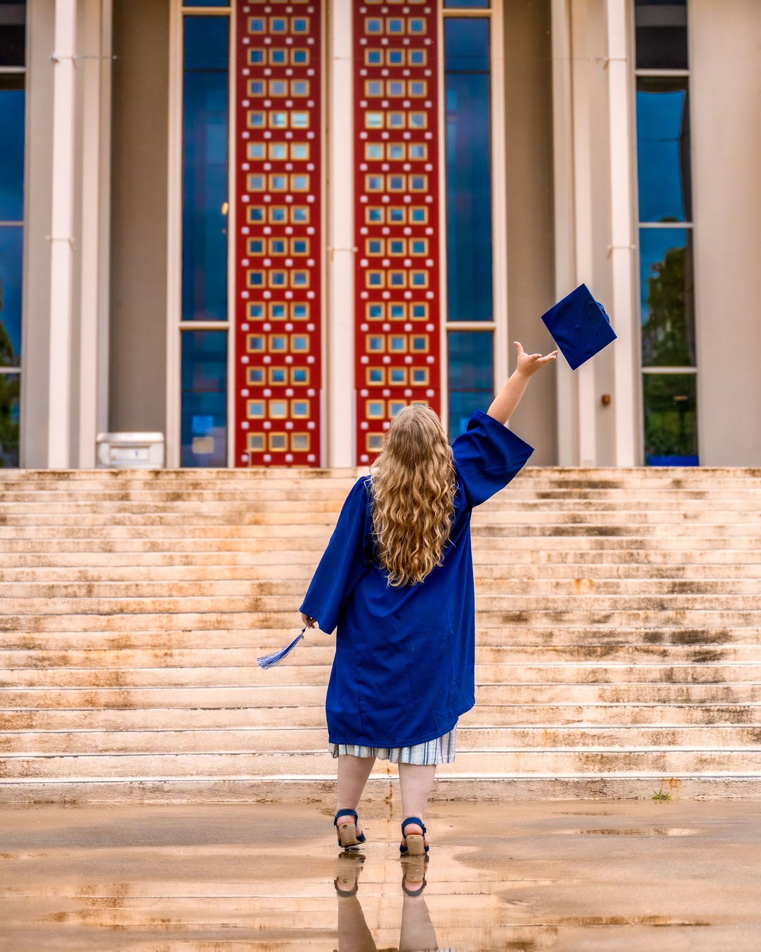 Graduate in blue cap and gown raising mortarboard toward grand building with red doors and stone steps.