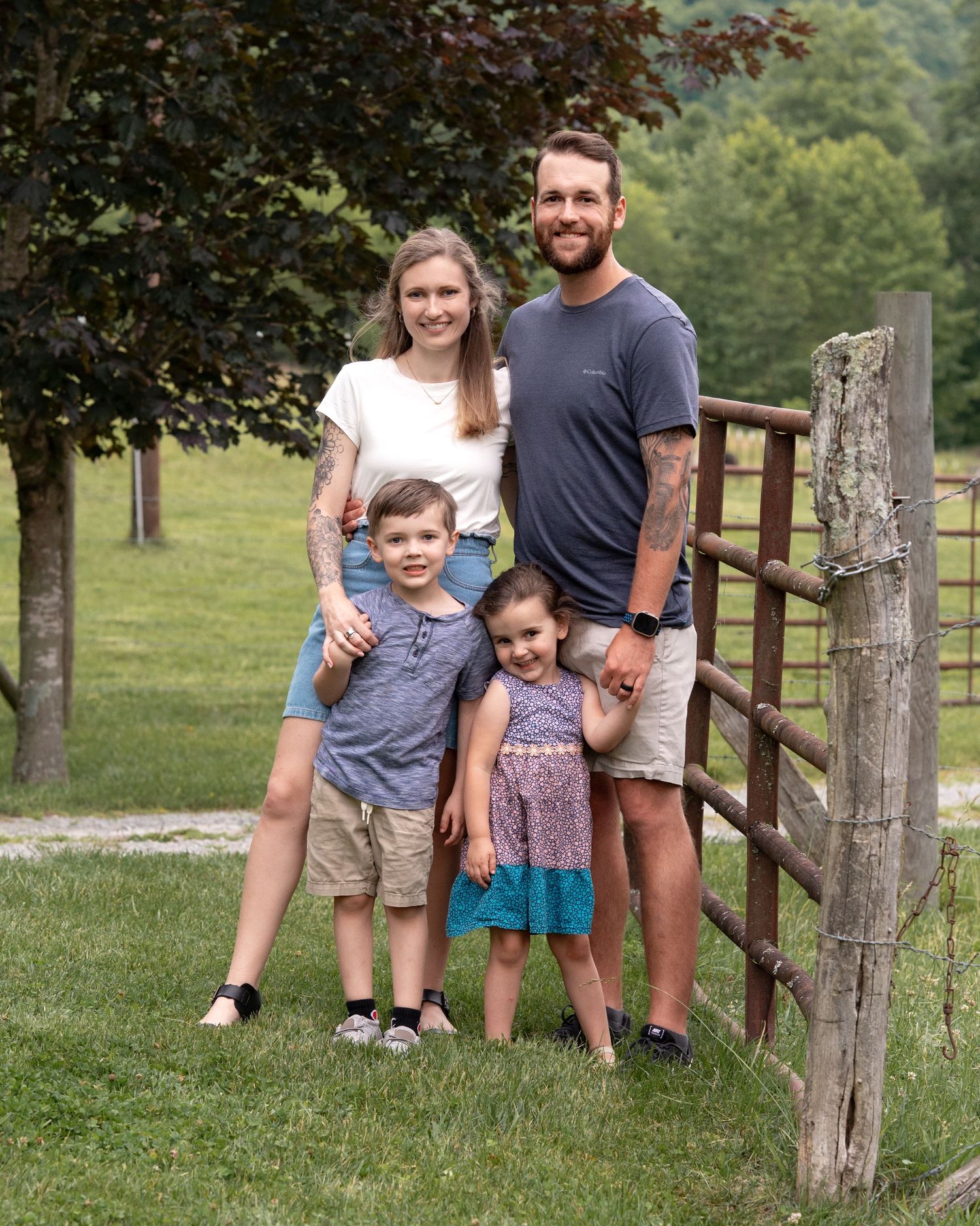 A happy family of four smiles together beside a wooden fence in a lush green countryside setting.