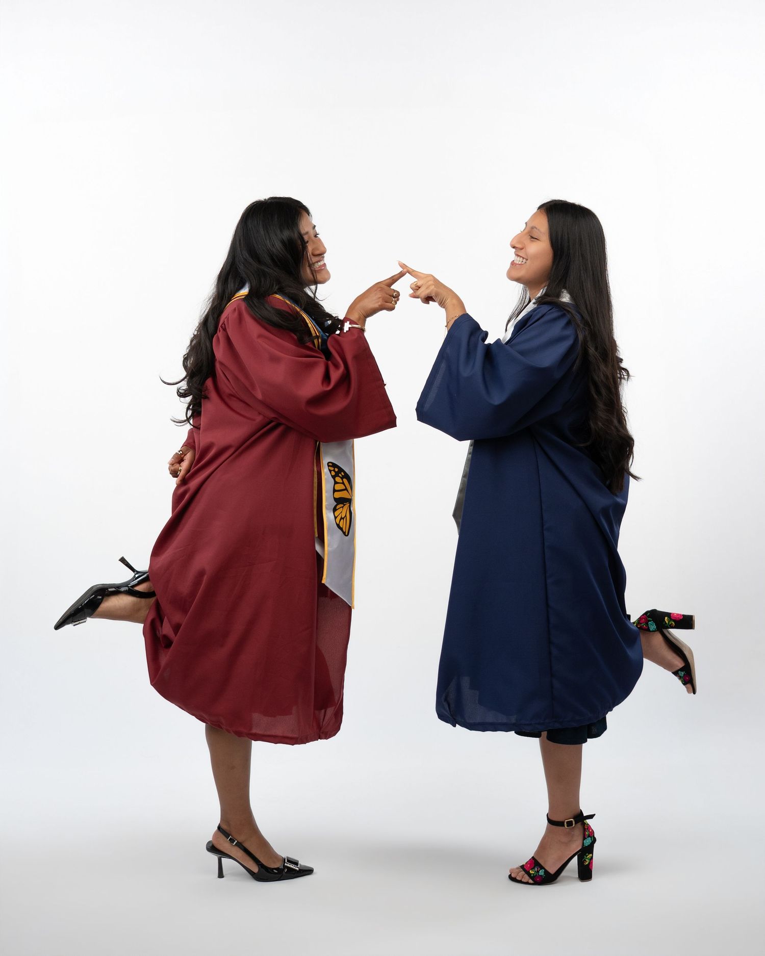 Two female graduates in maroon and navy gowns pose playfully pointing at each other in studio.