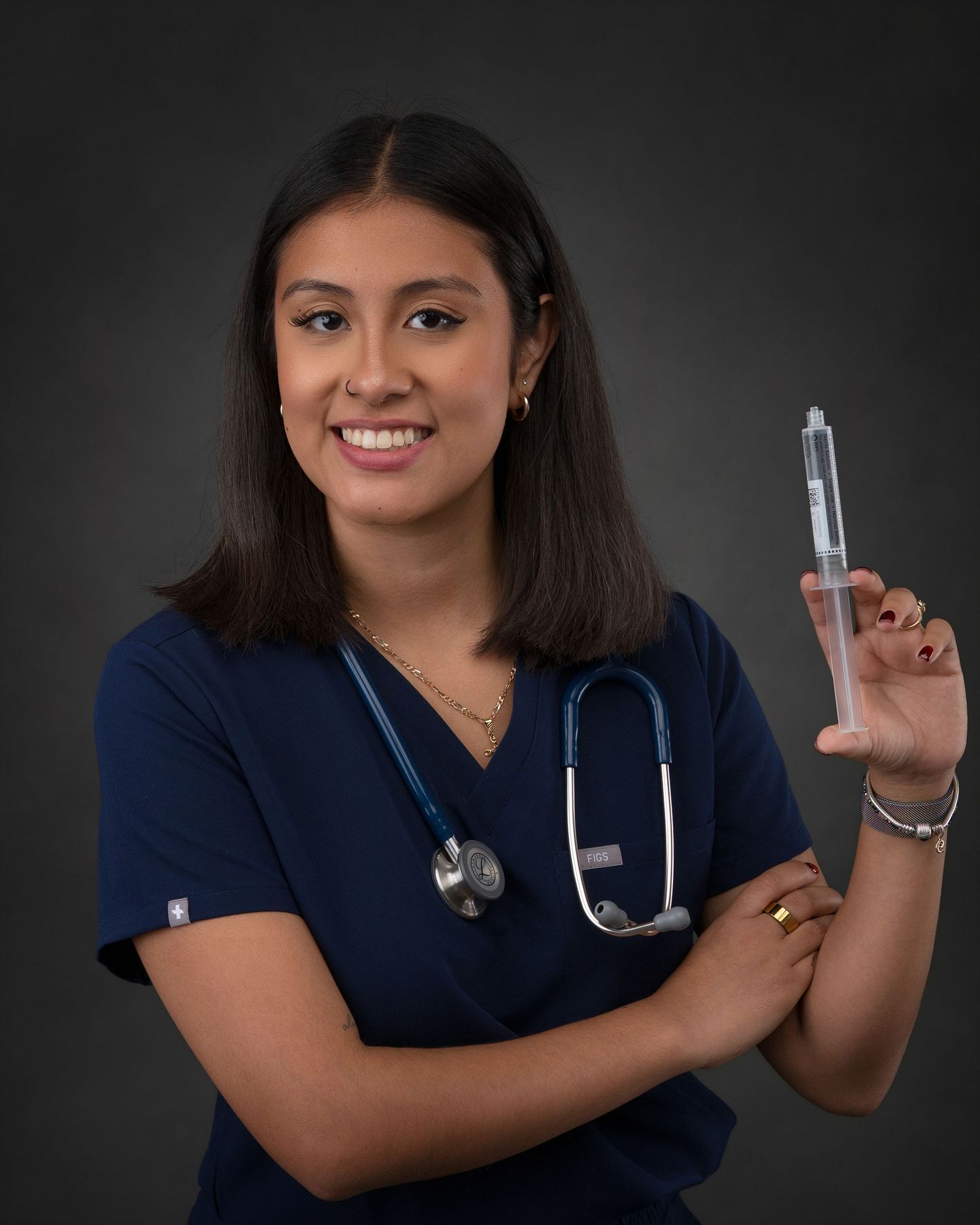 Smiling nurse in navy scrubs holds large syringe with stethoscope in studio portrait.