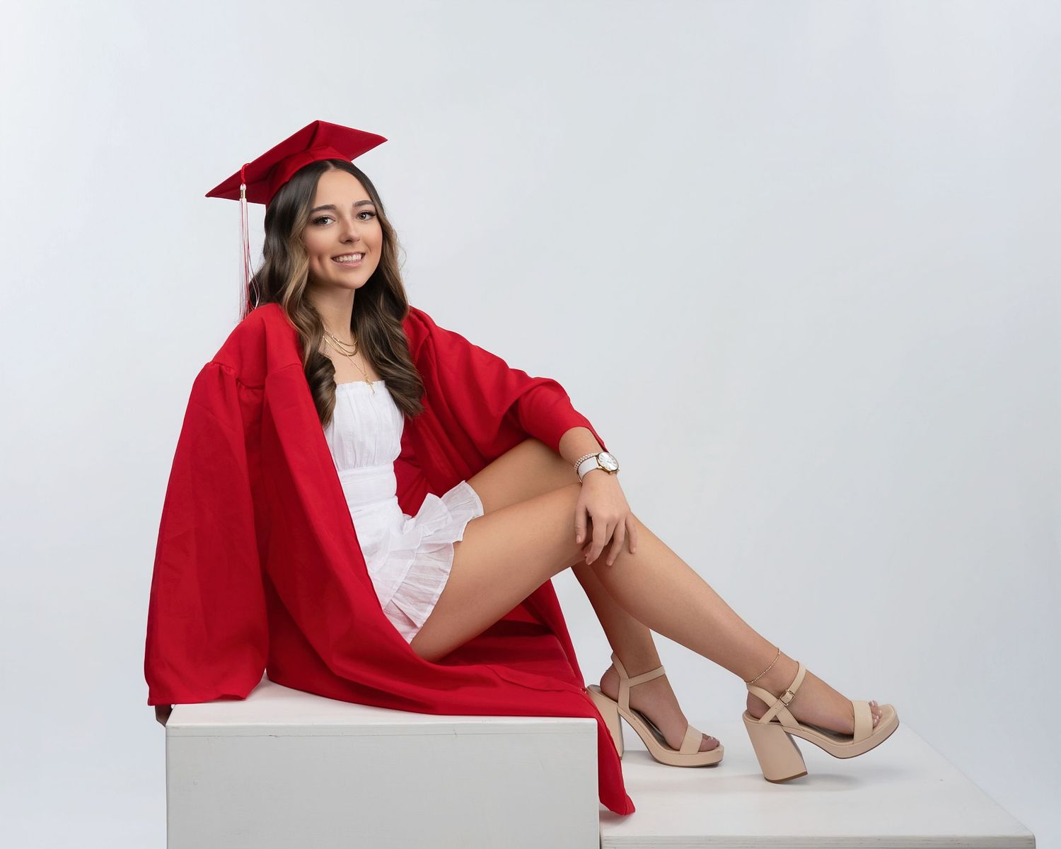 Smiling graduate in red cap and gown sitting on white cube wearing white dress and heels.