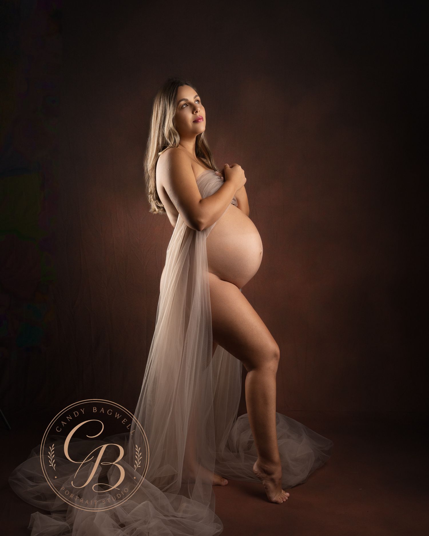 Pregnant woman in sheer white draped gown poses elegantly in a professional maternity portrait studio session.