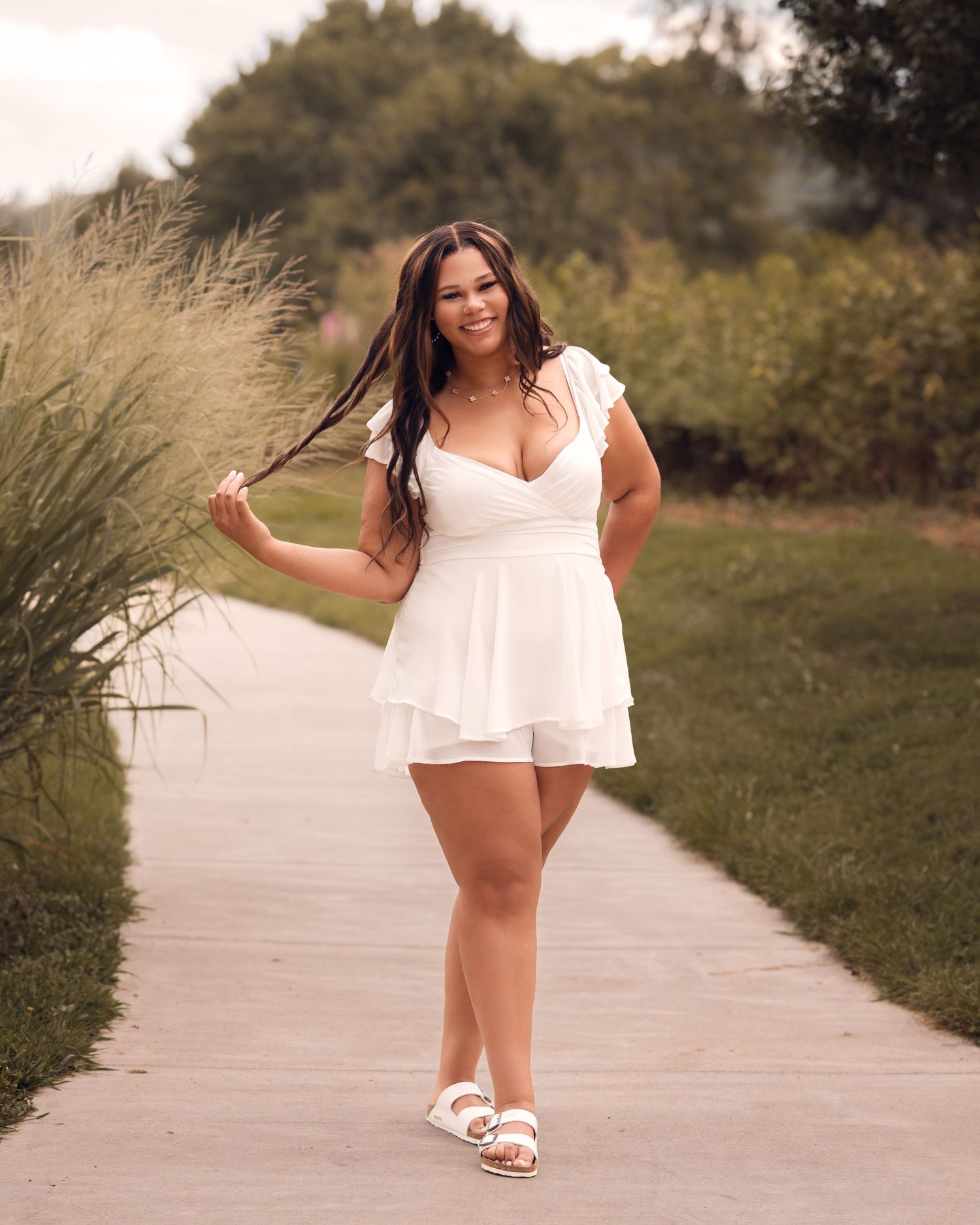 Smiling young woman in white romper walks along outdoor pathway surrounded by lush greenery.