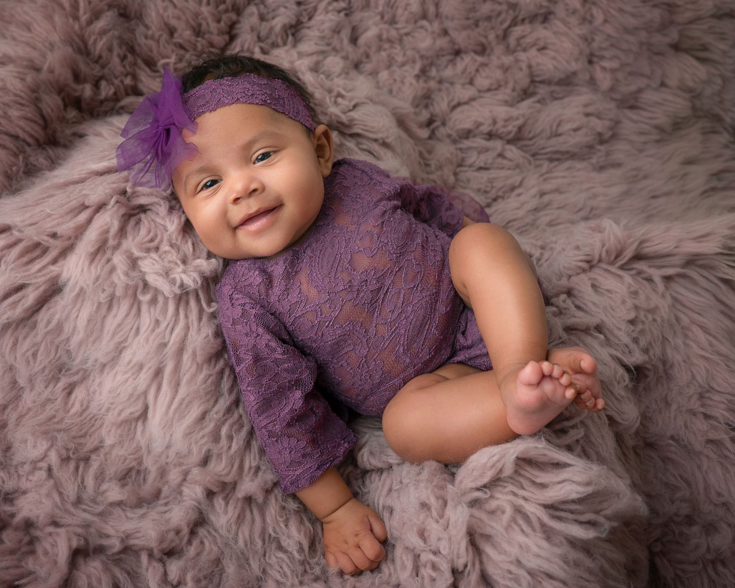 Smiling baby girl in purple romper and floral headband lies on fluffy lavender fur rug.
