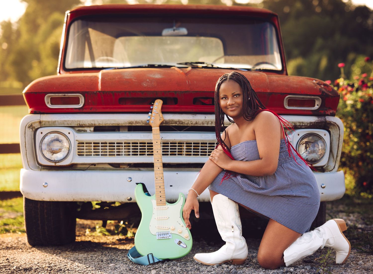 A smiling young woman in a dress poses with a green guitar beside a vintage red truck outdoors.