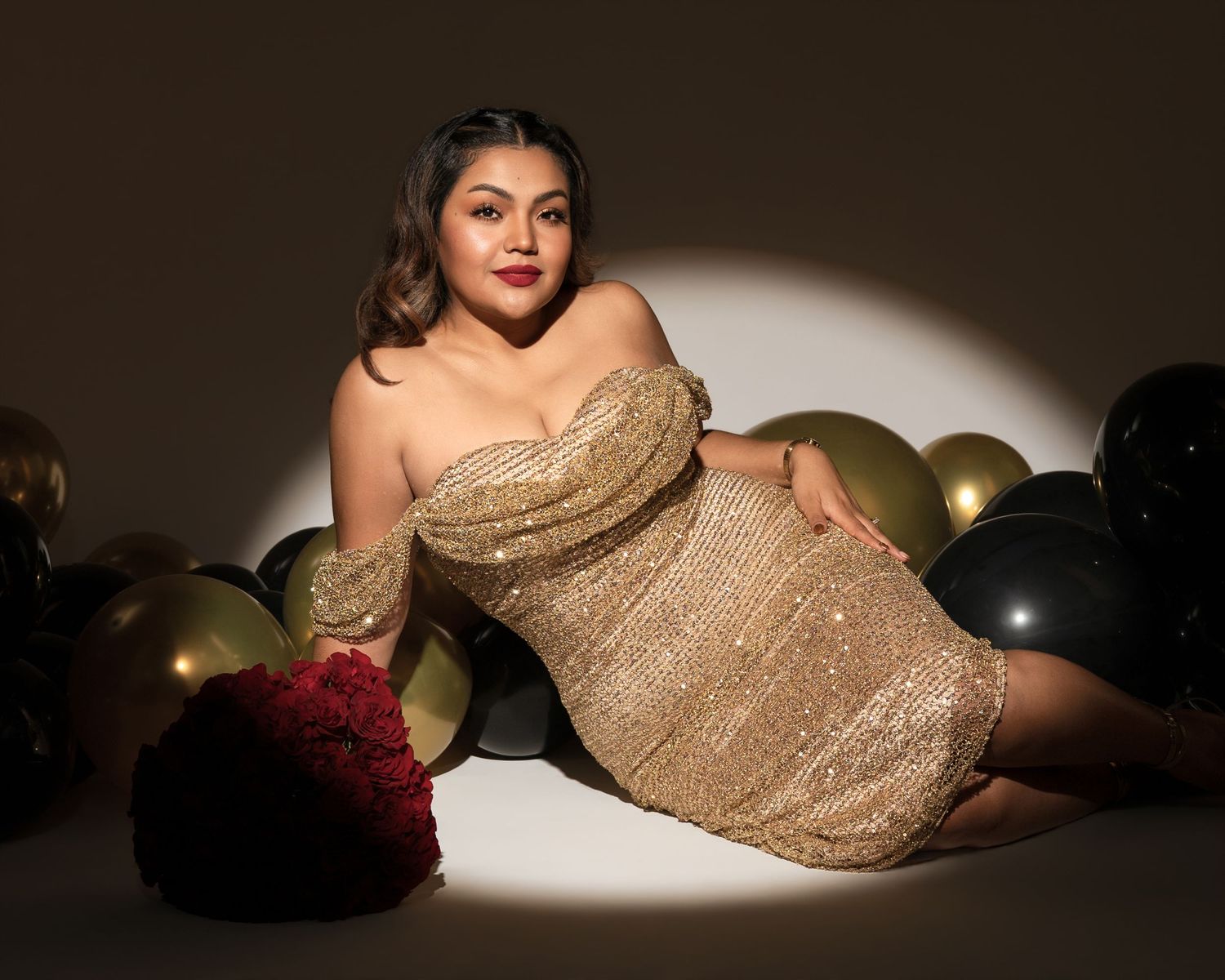 Woman in a sparkling gold off-the-shoulder sequin dress reclining on a spotlit floor among black and gold balloons beside red roses.