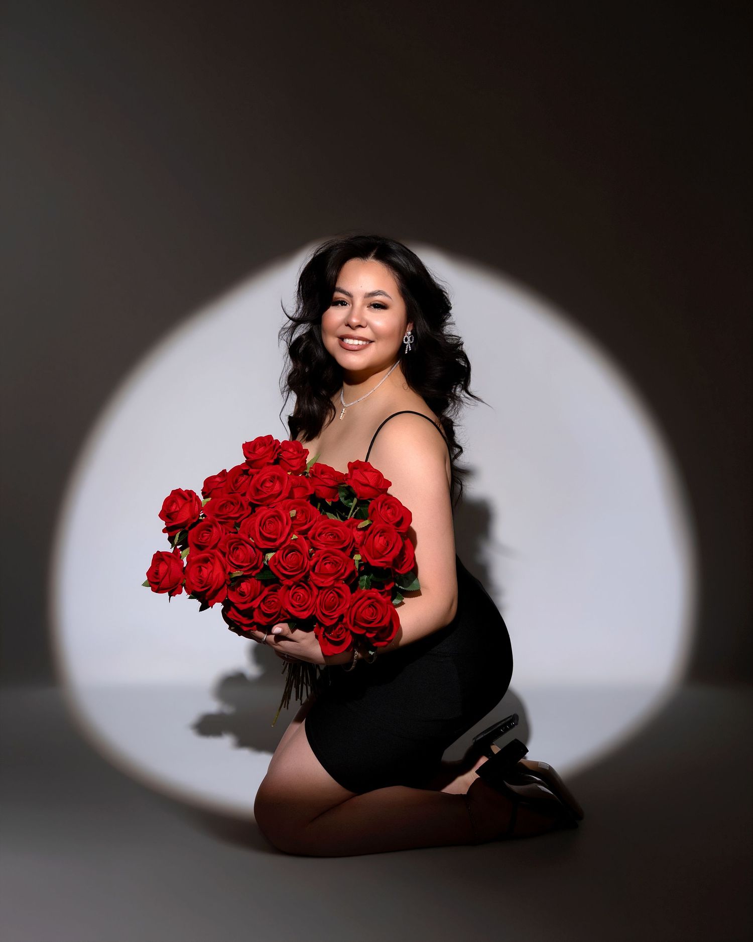 Woman in black dress holding large red roses bouquet while kneeling in circular spotlight studio.
